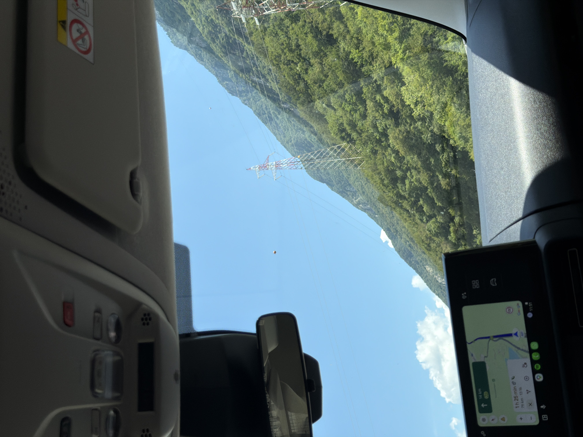 A view from inside a car shows a bright, sunny day with a clear blue sky. Mountains covered in lush green trees rise steeply to the right, with power transmission towers visible on their slopes. A rearview mirror is attached to the windshield in the foreground. Below the mirror, a dashboard-mounted screen displays a GPS navigation interface with a map and driving directions. The dashboard itself is dark and textured.