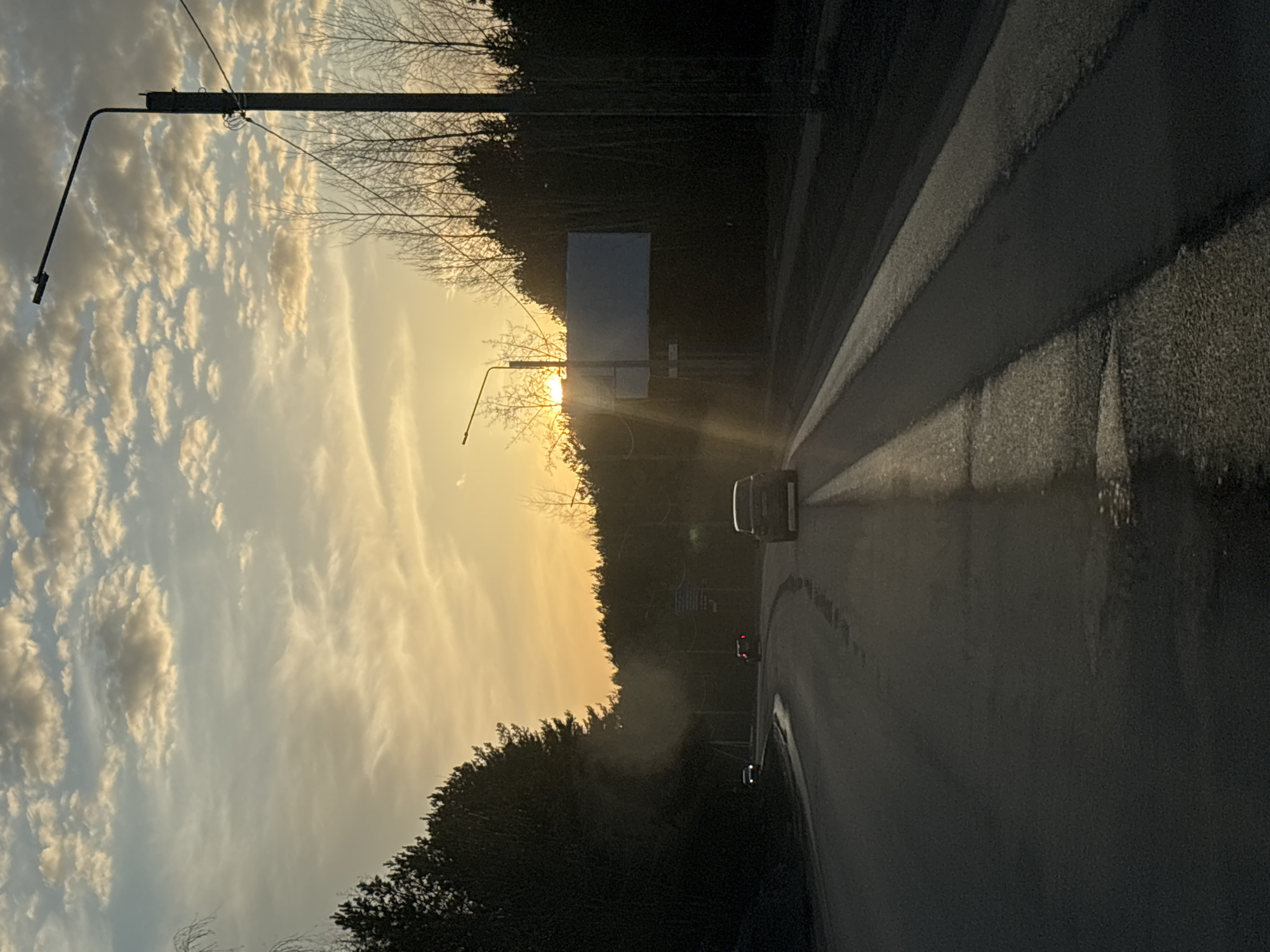 The image depicts a road scene in what appears to be the city of Chaikovsky, Russia, captured during sunset. The road curves gently to the right, with a few vehicles visible. A large sign is visible, and trees line both sides of the road, obscuring the view. A street lamp stands on the left. The sky is filled with clouds illuminated by the setting sun.