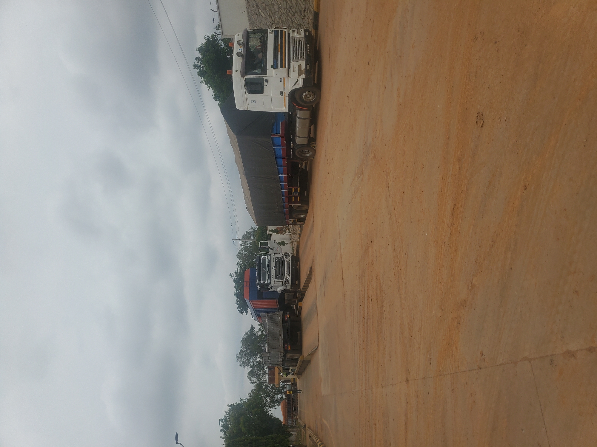 In Gwagwada, Nigeria, on a cloudy day, two large trucks are positioned on truck scales on a dirt road. The truck on the left is a dump truck with a blue and orange container. The truck on the right is a semi-trailer truck with a black tarp covering its cargo. The truck scales are elevated, with ramps leading up to them. A person wearing a high-visibility vest is standing near the truck on the left, possibly overseeing the weighing process. The environment appears to be an industrial or transportation hub, with trees and buildings visible in the background. The ground is covered in reddish-brown dirt, suggesting it may be a dry or arid region.