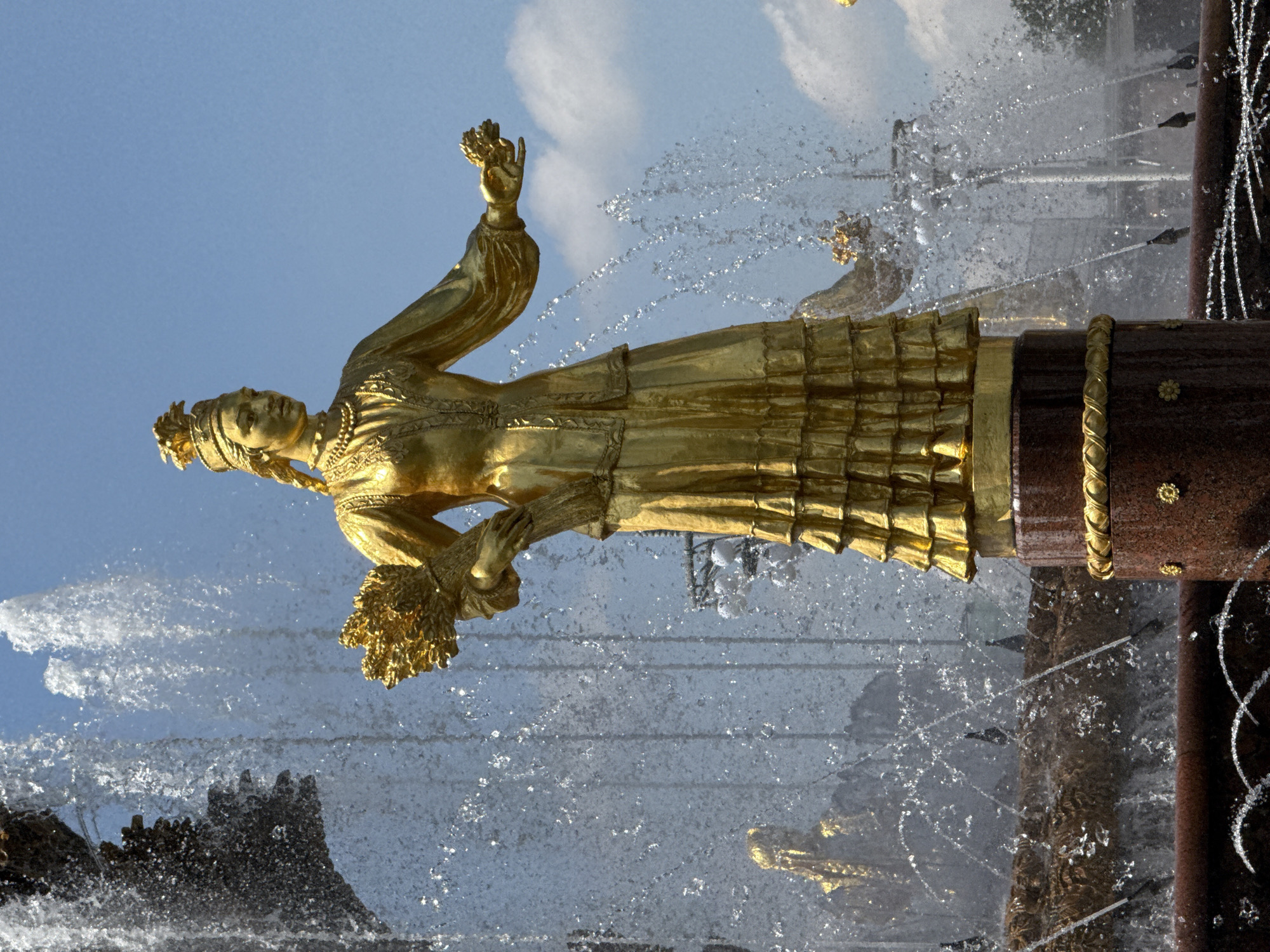 A golden statue of a woman stands on a fountain in Moscow, Russia. She is adorned in a traditional Russian dress and is holding a bouquet of flowers in one hand while raising the other. Water spouts around the statue, contrasting with the blue sky and white clouds in the background. The fountain structure is constructed of dark stone with gold accents. The statue appears to be one of the Golden Statues of the Friendship of Nations Fountain.