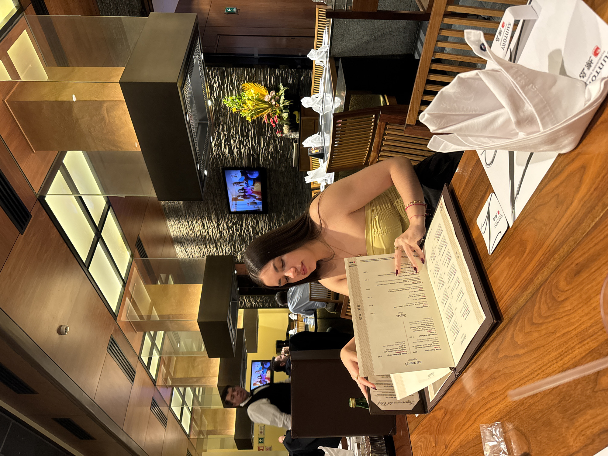 The image shows a woman reading a menu at a restaurant. She is wearing a yellow top and has long dark hair. The menu is in Spanish and English, and the Suntory logo is visible on nearby napkins. A waiter in a black vest and white shirt stands nearby. The restaurant has a modern design with wood paneling and glass accents. A television is visible in the background. Based on the Suntory branding, it is likely taken at the Suntory restaurant, a teppanyaki restaurant, which is in the city of Puebla, Mexico.