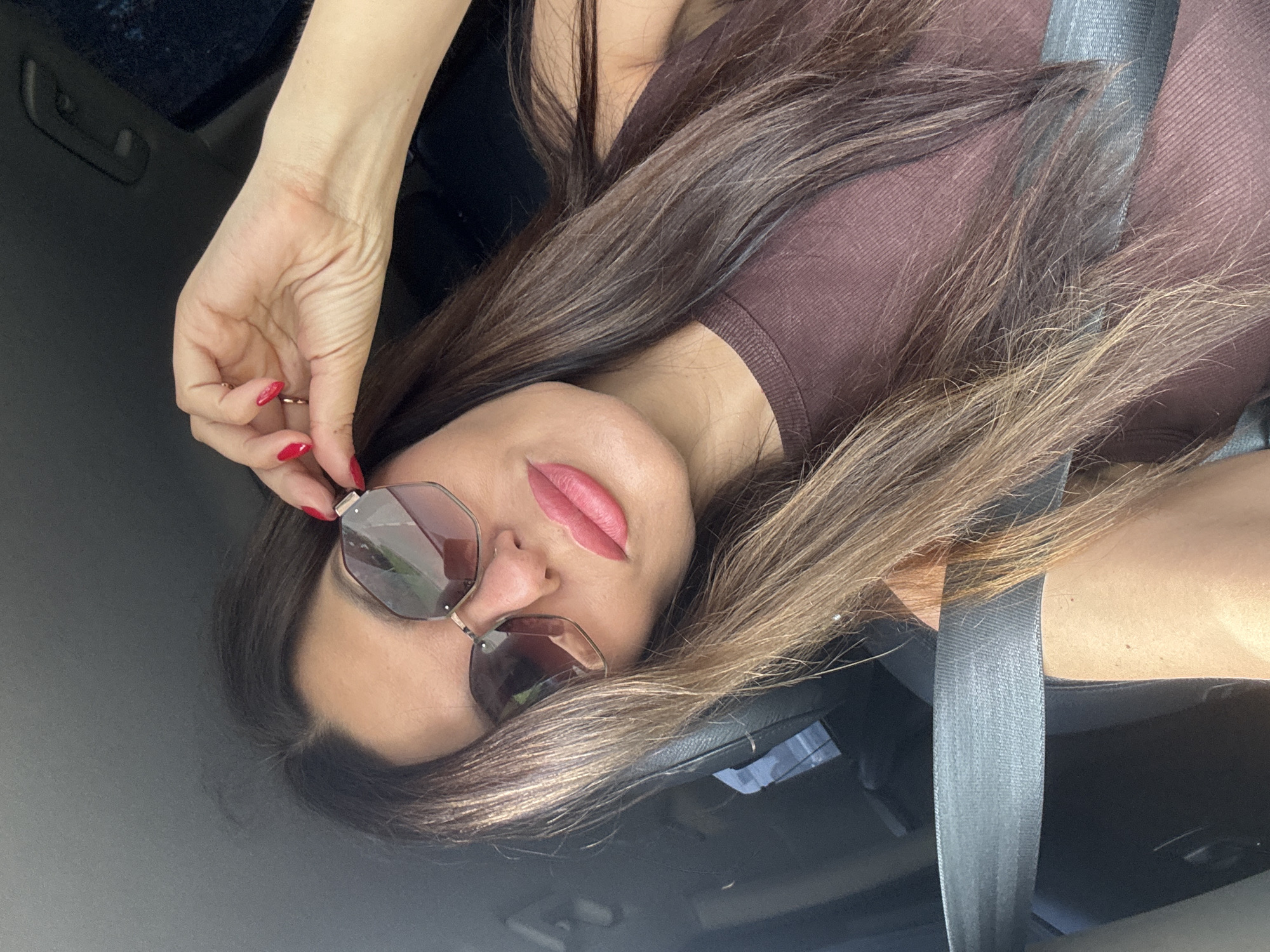 A woman is sitting in a car, looking at the camera. She is wearing sunglasses and a brown top. Her hand is on her sunglasses. The seatbelt is visible. The background is dark.