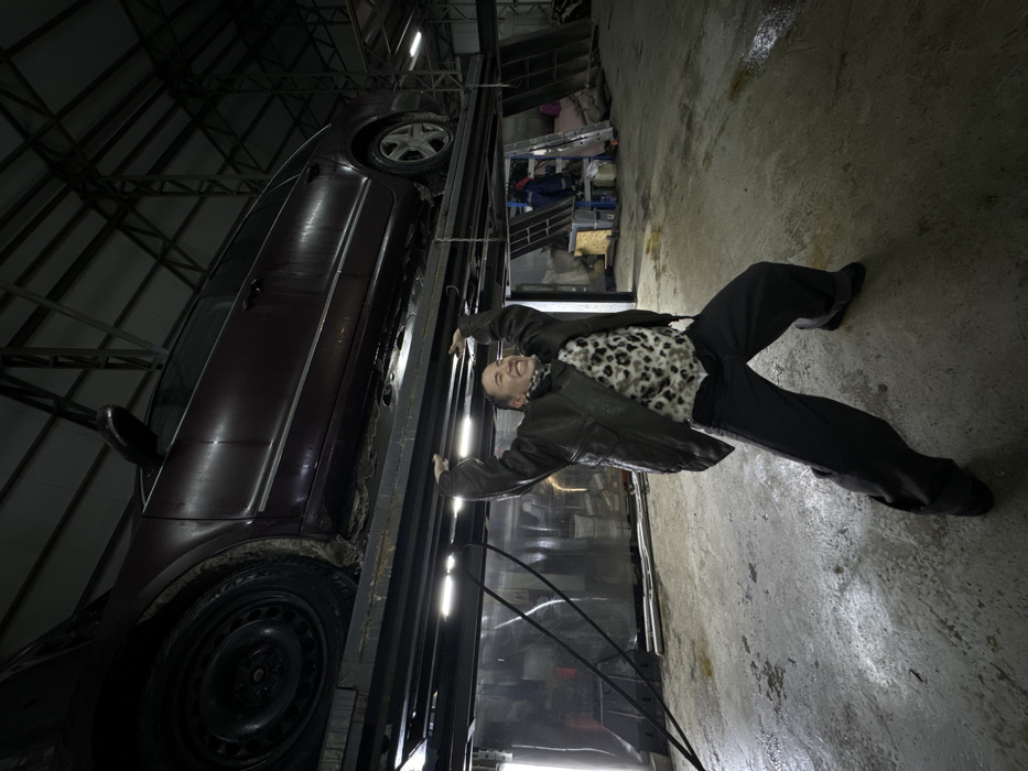 A man in a black jacket and pants with a leopard print shirt laughs while seemingly holding up a maroon car on a lift in a garage. The man has his arms outstretched and is positioned between the car and a shiny, reflective surface. The garage appears cluttered with various items visible in the background. The photo is taken from an unusual angle, giving the impression that the car and lift are tilted precariously.