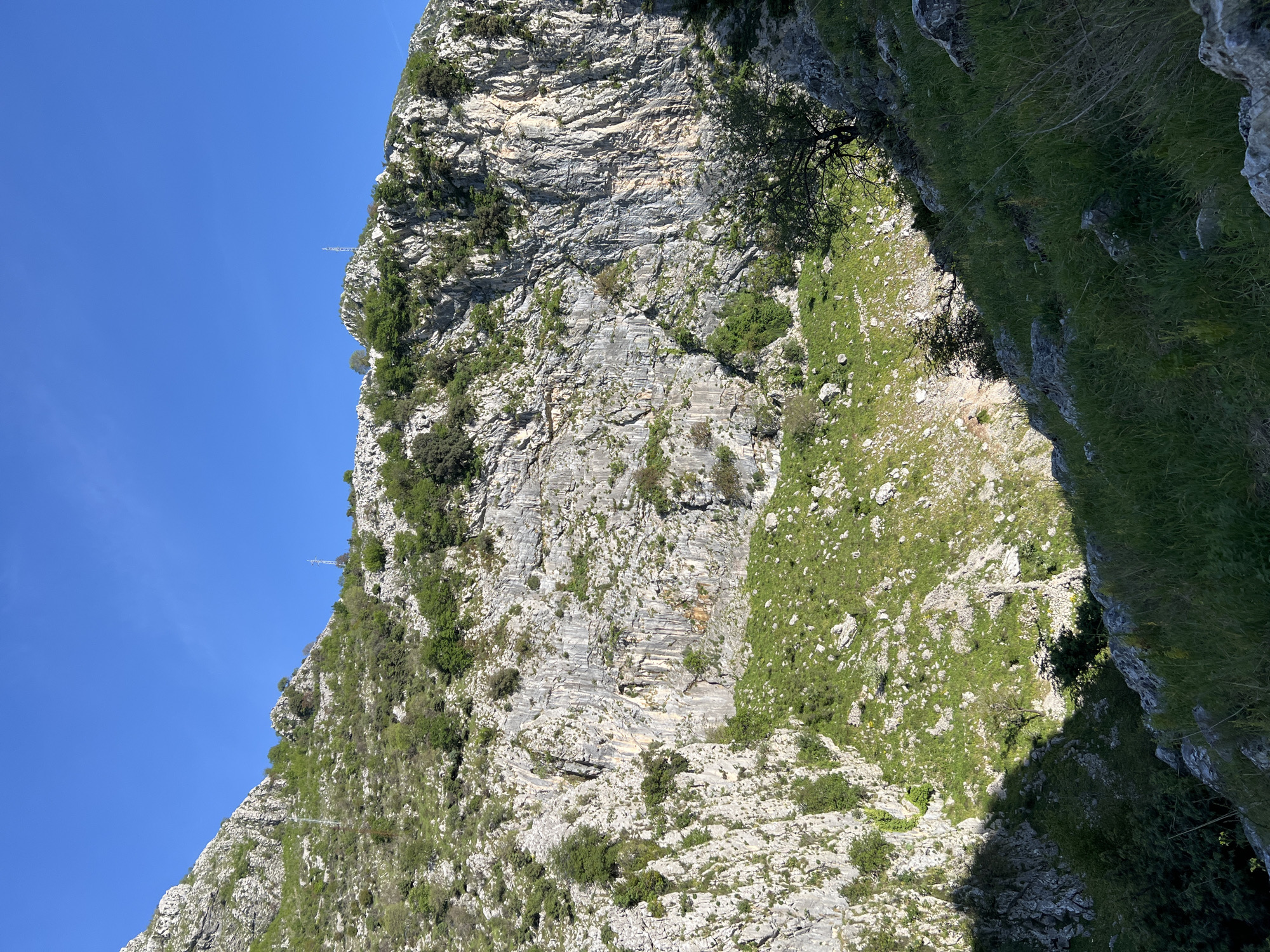 The image displays a rugged mountainous landscape under a clear blue sky. A massive, steep rock face, characterized by light grey, horizontally layered rock formations, dominates the right side of the frame. This cliff is interspersed with patches of green vegetation, including shrubs and small trees, growing in its crevices and on ledges. High on the mountain's crest, against the bright blue sky, two slender lattice structures, likely power line pylons or communication towers, are faintly visible in the distance.
To the left of the prominent rock face, a steep, verdant slope descends. This grassy area is dotted with scattered rocks and sparse bushes. A significant portion of the slope is cast in deep shadow, creating a strong contrast with the sunlit areas and suggesting a deep ravine or a shadow from an adjacent elevation. A narrow, light-colored path or ancient stone terrace can be seen winding horizontally across the grassy slope in the mid-ground.
The scene is captured on a clear, sunny day, with strong sunlight and distinct shadows. No specific activity or individuals are present. The formidable rock formations and steep terrain are characteristic of the mountainous regions around Kotor, Montenegro. There is no visible text in the image.