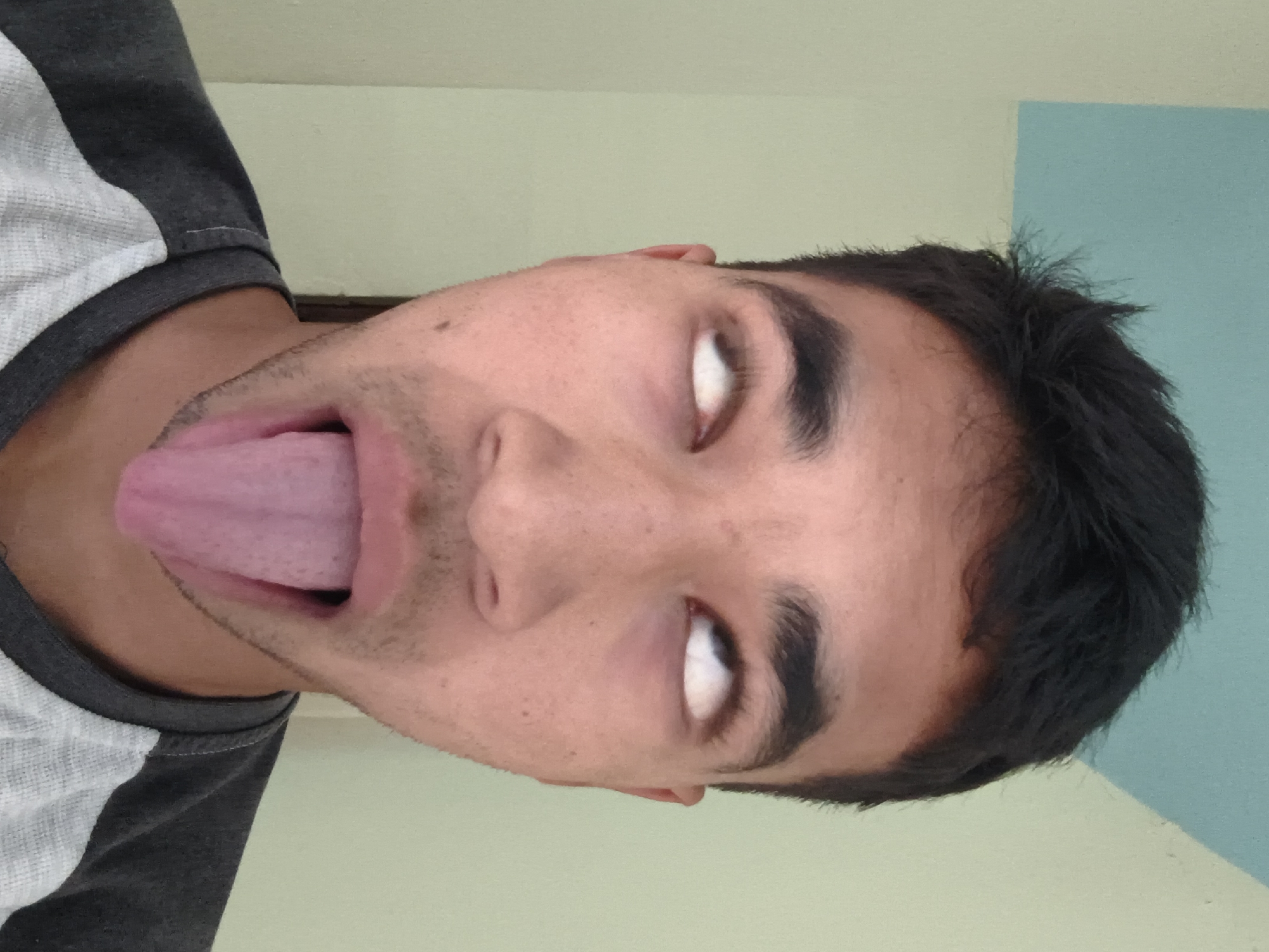 The image shows a close-up, head-on view of a young man's face. His eyes are rolled back, showing only the whites, and his mouth is open with his tongue protruding. He is wearing a gray and white t-shirt. The background consists of a wall painted in two colors: pale green on the left and a light blue rectangular area on the top right. There are no other people or objects visible in the frame, and there is no discernible information about the location being Haini, India. The lighting suggests an indoor environment with natural light coming from the front.