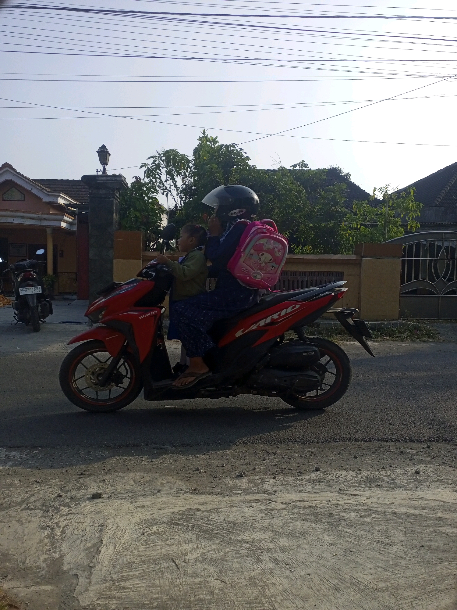 Two individuals, a child and an adult, are riding a red Honda motorcycle on a paved road in the city of Ngasem, Indonesia. The adult is wearing a black helmet and the child is wearing a pink backpack adorned with a cartoon character. The motorcycle has a black seat and black tires with red accents. In the background, a house with an orange facade and a dark roof is visible. Trees and power lines are also present in the scene. The road surface is asphalt with some areas of concrete. The lighting suggests it is daytime, possibly late afternoon, with a clear sky.
