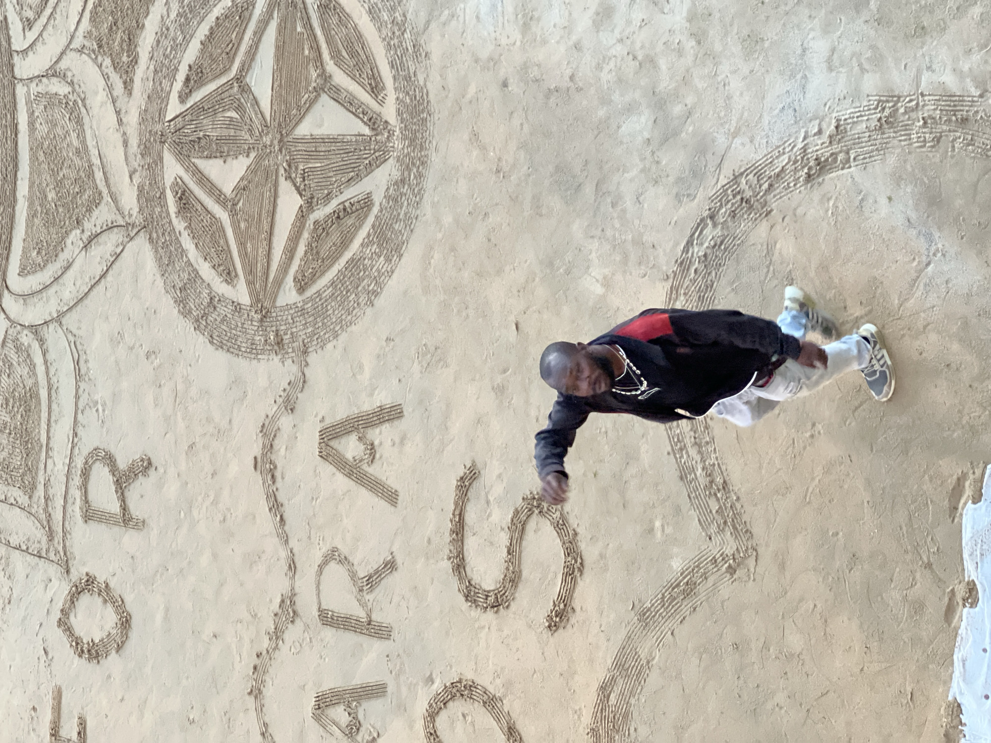 An aerial, high-angle view shows a man in casual clothing on a sandy beach in San Sebastián, Spain. He is wearing a black jacket with red accents on the sleeves, light-colored pants, and athletic shoes. The man is walking and gesturing with his right hand. In front of him, etched into the sand, are large letters that appear to spell "FOR AMARA OS" and decorative patterns, including a compass rose and a heart shape. The beach is wet and the sand shows textures from the etching process.