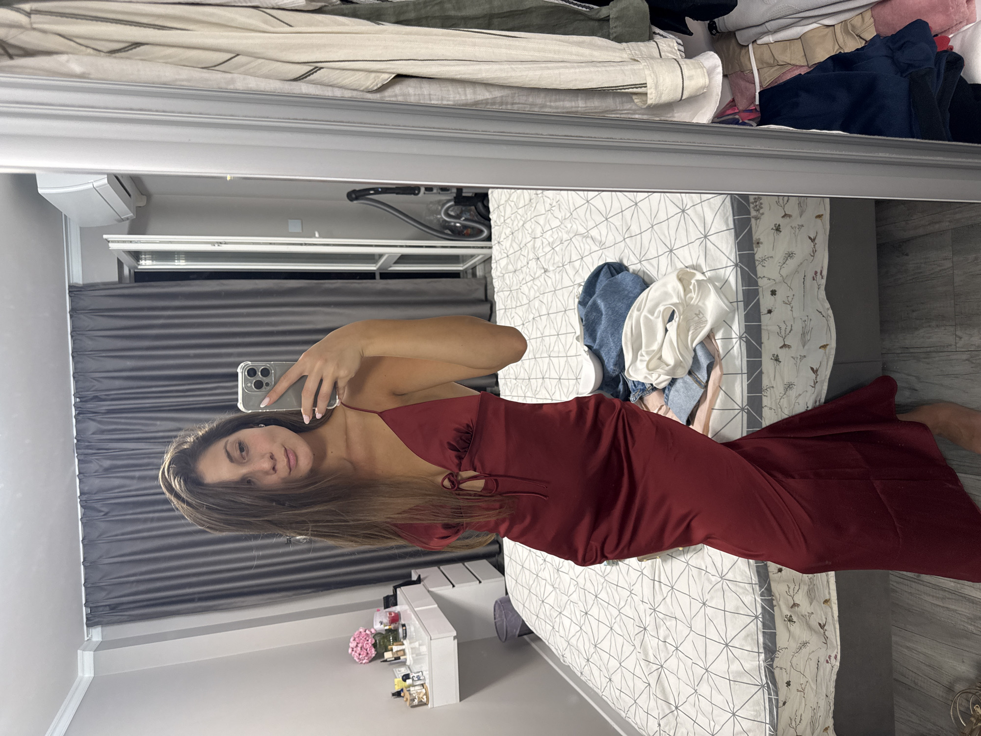 A young woman stands in a bedroom, taking a mirror selfie. She is wearing a long, form-fitting, dark red satin dress with spaghetti straps and a tie detail at the bust. Her long, brown hair is down, and she is looking at her phone. The bedroom appears to be somewhat cluttered. A bed is visible behind her, covered with a white and grey patterned duvet, and several articles of clothing are draped over it. To the left of the bed is a white vanity table with various cosmetic items and a pink bouquet of flowers. A trash can is next to the vanity. To the right of the woman, a full-length mirror reflects a portion of the room, including clothes hanging on a rack. The environment suggests a residential setting, likely a bedroom in an apartment. The image does not contain specific visual cues to pinpoint the location to Burgas, Bulgaria, or any other specific city.