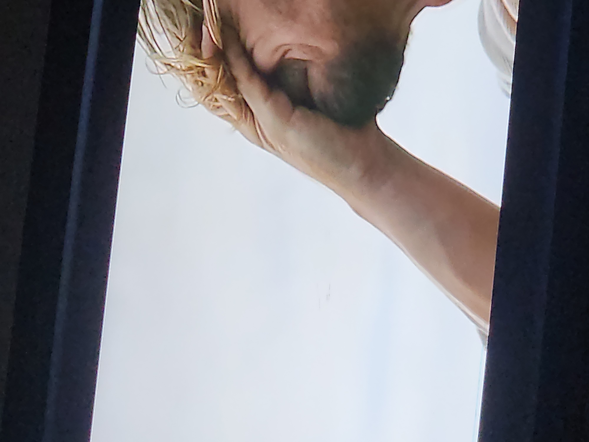 The image shows a close-up, low-angle shot of a person's arm and head, with their hand covering their face. The person has light-colored, messy hair. They are wearing a light-colored garment, possibly a t-shirt. The background is a bright, overcast sky. The bottom of the image shows a dark, rectangular frame, suggesting the image was captured through a screen or a window with a dark border. There is no indication of the city of Los Robles, Venezuela, or any other specific location within the image.