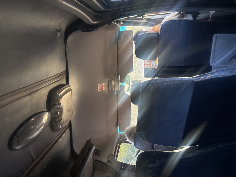 The image depicts the interior of a vehicle, likely a minibus or shuttle, in Partskhanakanevi, Georgia, viewed from a rear passenger seat looking towards the front. The light gray headliner includes an oval chrome-finished light fixture and a beige plastic overhead control unit with red indicator lights. Two beige sun visors are visible, one positioned centrally and another angled to the right. A red and white circular "No Smoking" sign, displaying a crossed-out cigarette, is affixed to the headliner above the right sun visor. The backs of several dark blue passenger seats are prominent in the foreground and midground. Sunlight intensely streams through the front windshield, creating significant glare and lens flares across the scene. The driver, identifiable by the back of a head with short gray hair, is visible in the front left seat. A second individual, with the back of their head and shoulder visible, occupies the front right passenger seat, partially obscured by the glare, with a hint of an orange garment. Through the windows, green foliage is discernible outside the vehicle.