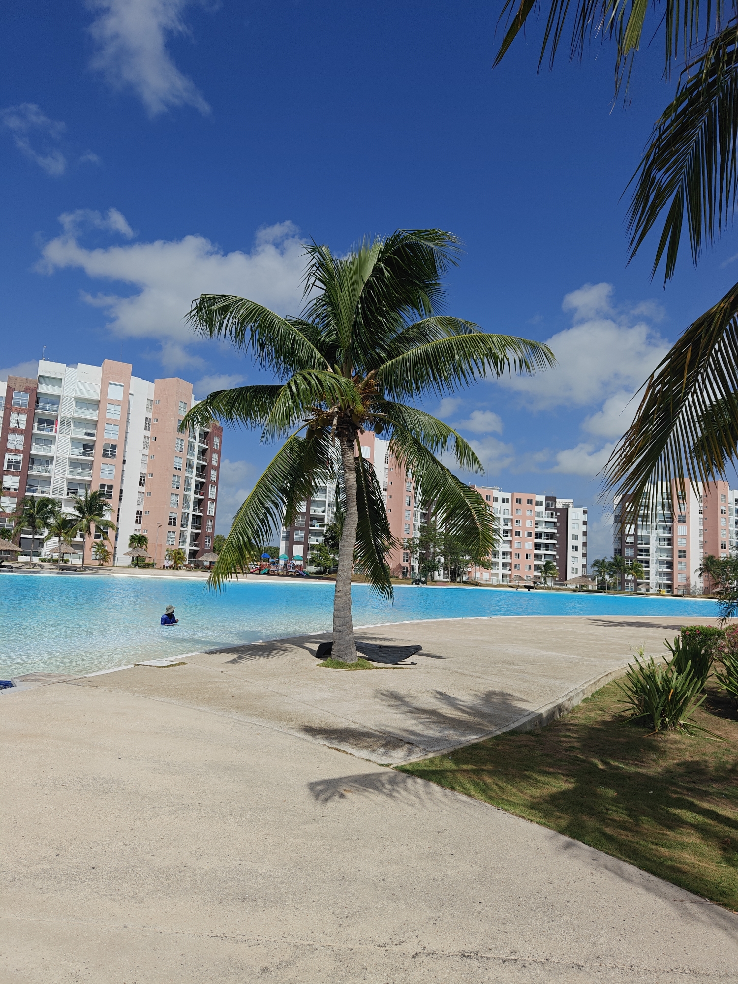 In an outdoor scene, a person wearing a blue shirt and a hat stands in a shallow, turquoise pool. Palm trees cast shadows on a concrete walkway and the surrounding grass. In the background, several tall, multi-story buildings with balconies and windows rise against a bright blue sky with scattered clouds. The buildings appear to be apartment complexes or hotels. The scene depicts a resort-like environment.