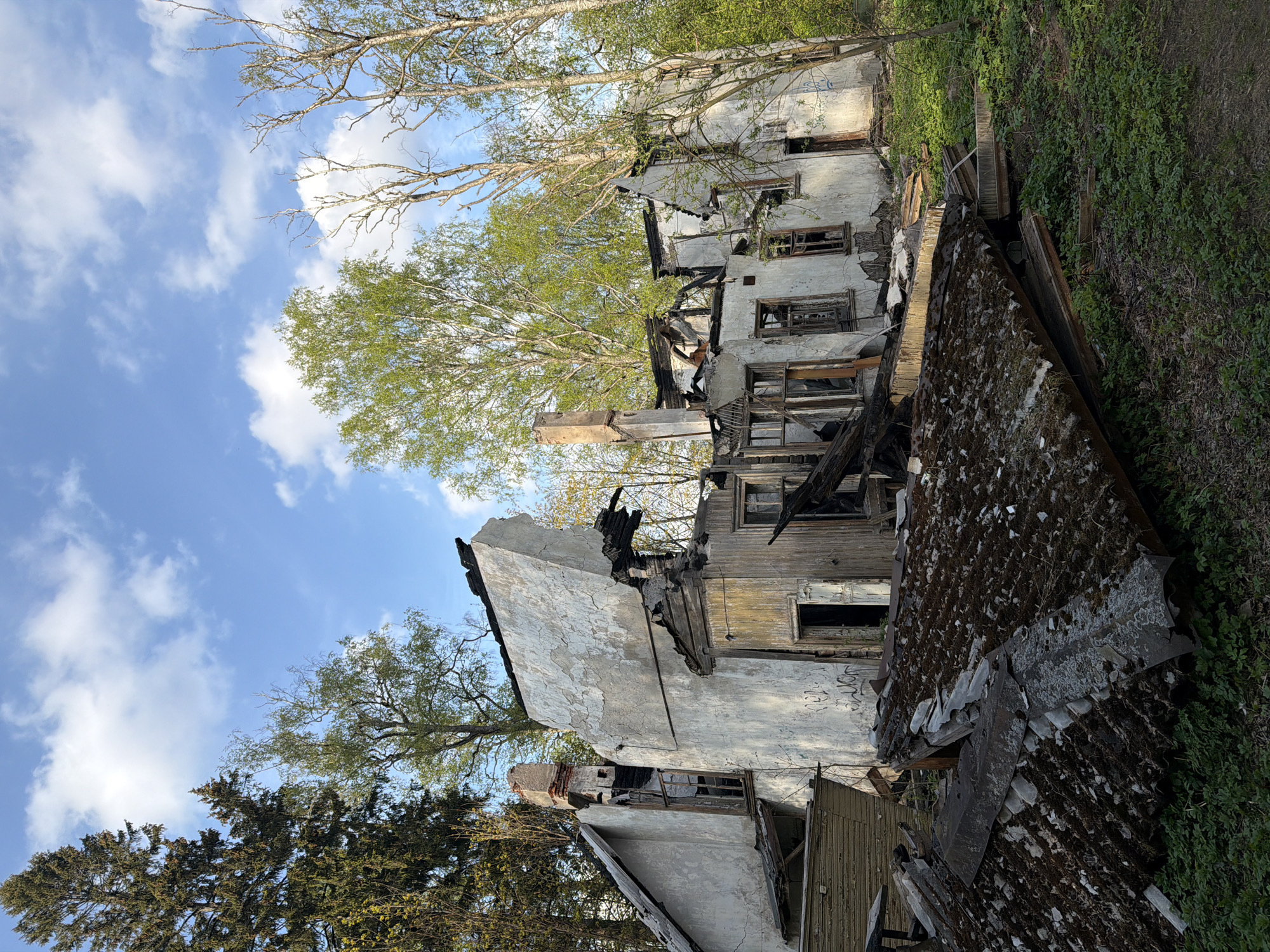 The image depicts the ruins of a building, possibly a house, in an overgrown environment. The building appears to be heavily damaged, possibly by fire, with charred remains visible. The walls are crumbling, and the roof is partially collapsed and covered with moss. Windows are broken or missing. The scene includes trees and vegetation encroaching upon the structure. There are no people visible in the image. The sky is visible with a mix of blue and clouds. There is no basis to suggest the image is in Tokkari-Lend, Russia.