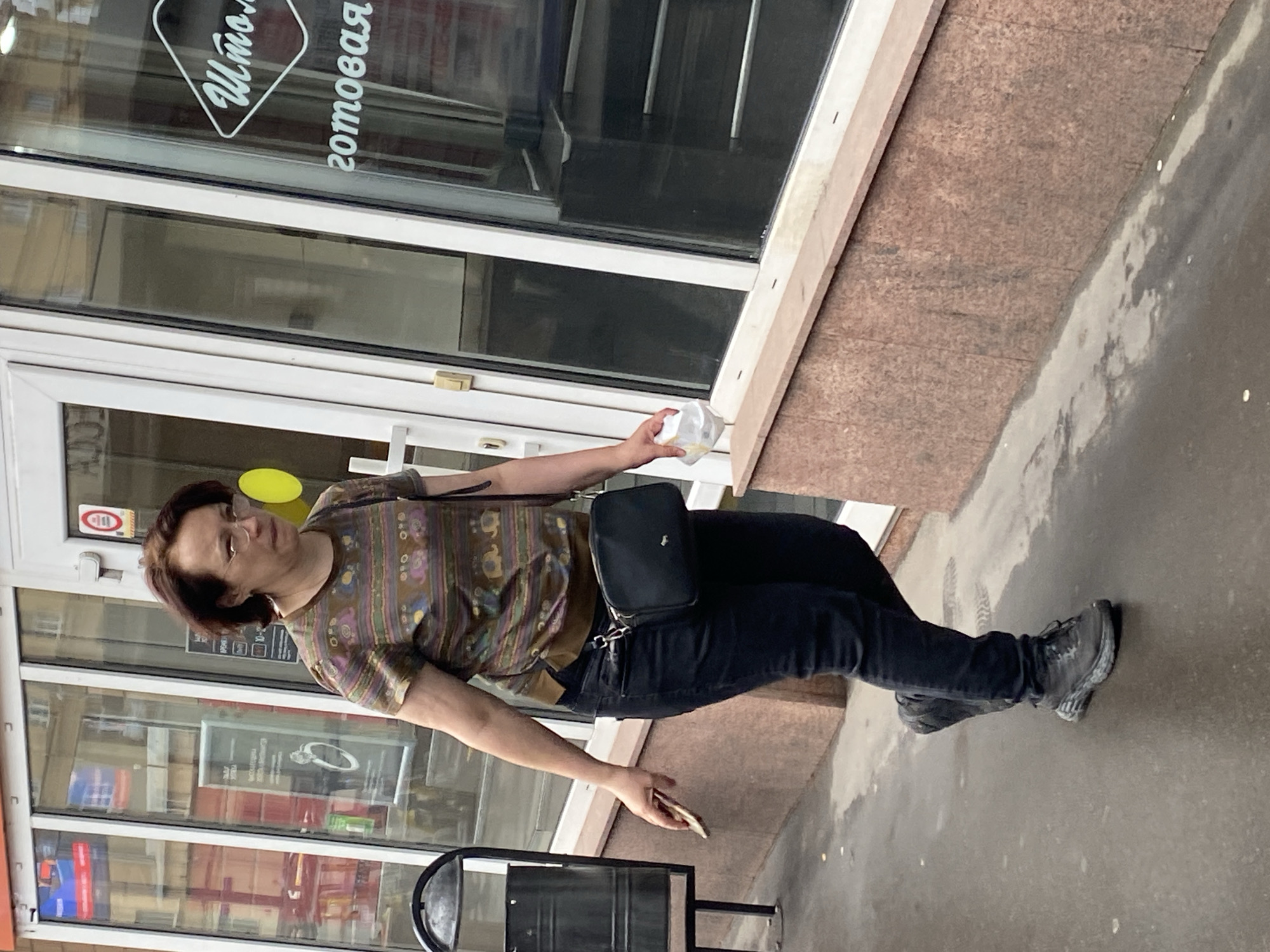 A woman with short, dark hair and glasses walks on a wet sidewalk in front of a building with large glass windows. She is wearing a patterned short-sleeved shirt, dark jeans, and dark sneakers. A black crossbody bag is slung across her body. She is holding a smartphone in her left hand and a white plastic bag in her right hand. The building appears to be a commercial establishment, with signs visible through the windows. A trash can is located to the left of the woman. The environment suggests an urban setting in Khimki, Russia, likely during overcast or rainy weather.