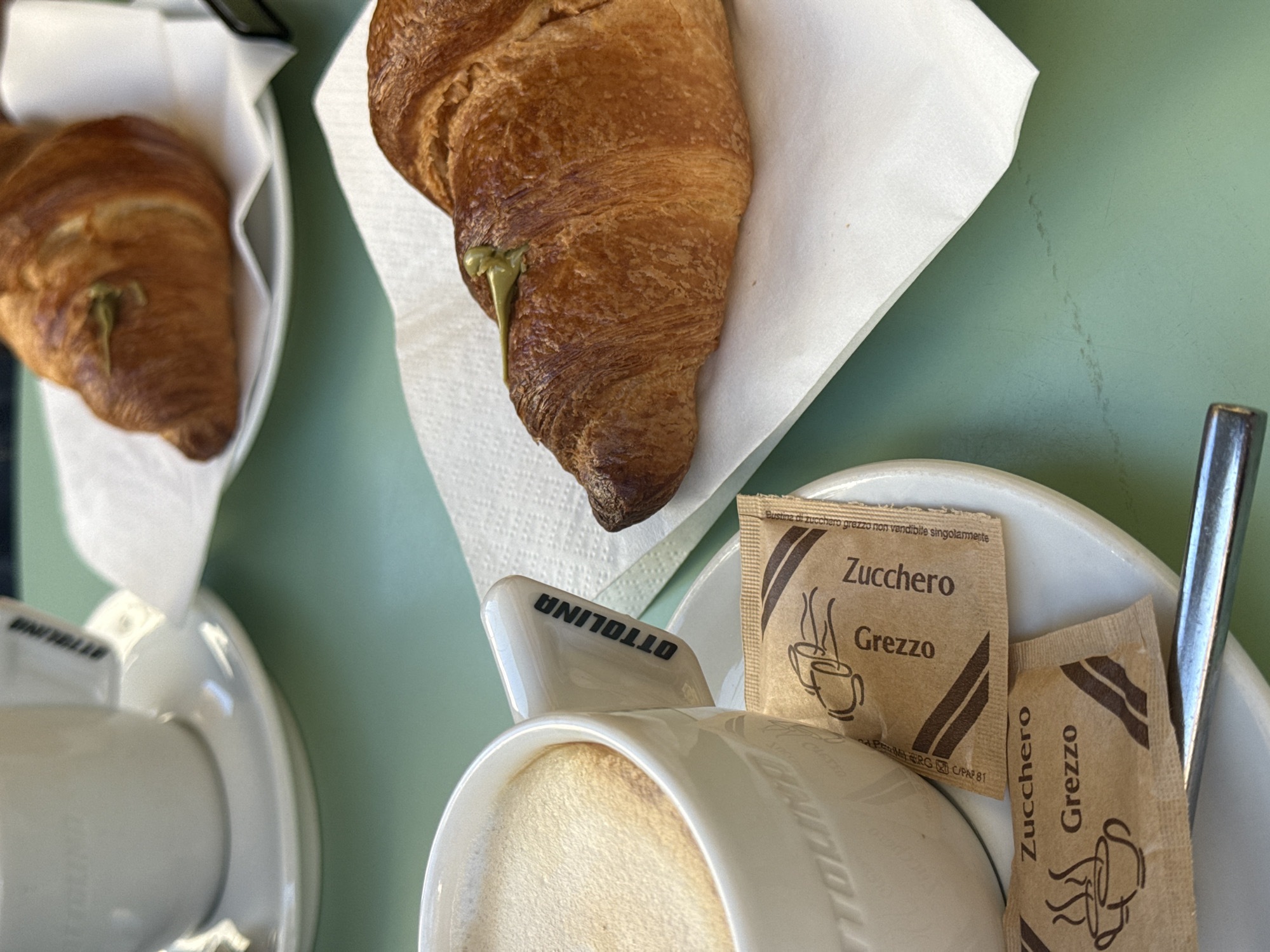 The image shows two pistachio croissants and two cups of coffee on a green table. Each croissant sits on a white paper napkin. Near one of the cups of coffee are two brown sugar packets labeled "Zucchero Grezzo" in Italian. The cup reads "OTTOLINA" and "BANYELER" indicating the location as a Milan, Italy establishment. A metal spoon rests on the saucer of the coffee cup.