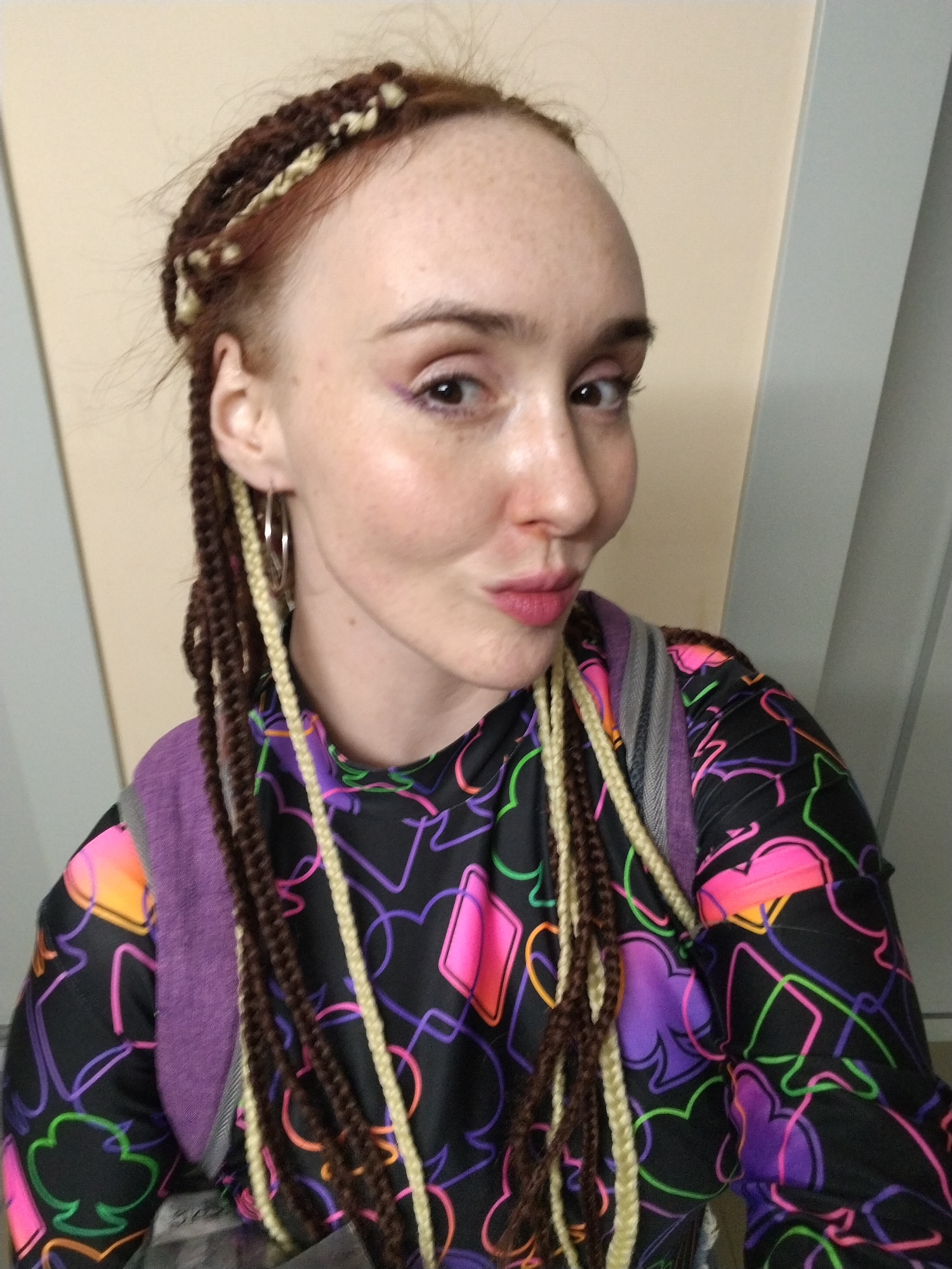 A fair-skinned woman with red hair and freckles poses indoors against a beige and white wall. She has her hair in braids of mixed dark brown and blonde hues. She wears silver hoop earrings and purple eyeliner. She wears a black long-sleeved shirt printed with neon playing card suits. She is wearing a gray and purple backpack. The location is not identifiable as the city of Izhevsk, Russia.