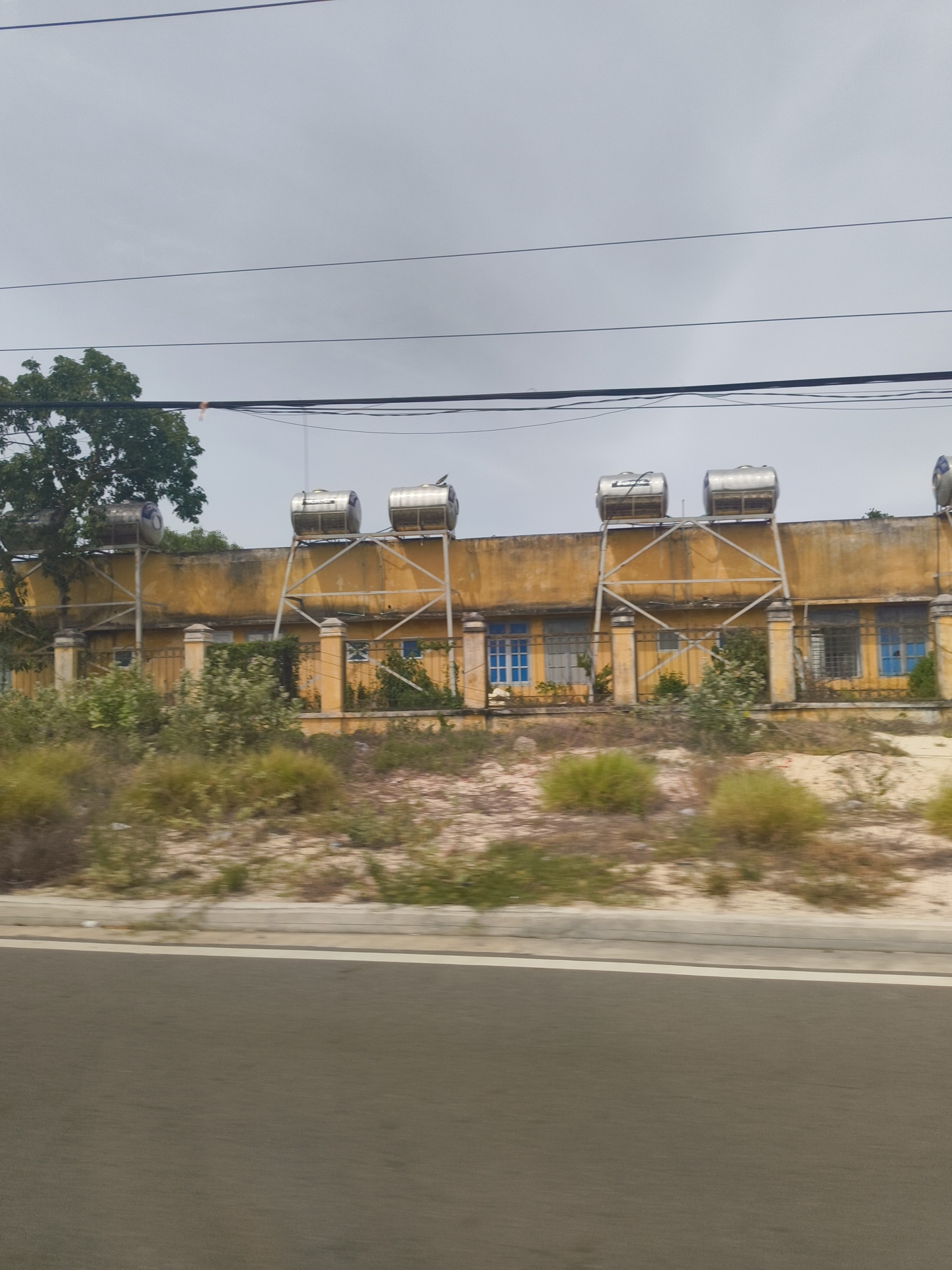 A long, one-story building with a yellow, weathered facade stretches across the frame, viewed from a slight side angle. The building has multiple windows, some with blue panes and others with barred sections. A low, yellow-capped fence runs in front of the building. Several large, cylindrical, silver water tanks are mounted on metal frames atop the roof of the building. To the left of the building, a large tree with dense green foliage is visible. The foreground is dominated by a grey road with a white stripe on its left edge, suggesting the photo was taken from a vehicle moving along the road. The ground in front of the building is covered in dry, sandy earth with sparse patches of dry grass and shrubbery. The sky above is overcast and pale grey, with several dark, horizontal power lines crossing the upper portion of the image. The scene appears to be in a rural or semi-urban area, possibly a school or administrative building, given the infrastructure. The location is identified as Bac Cam Ranh Ward, Vietnam.