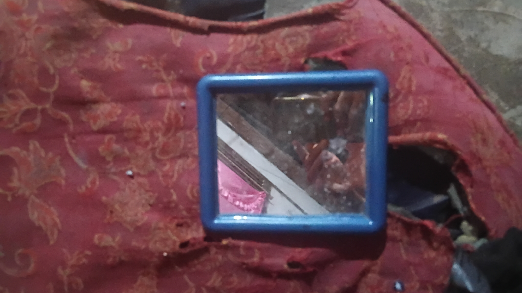 A small, blue-framed mirror rests on a damaged, red patterned mattress. The mattress appears old and worn, with visible tears and holes, revealing a darker interior. The mirror reflects a dim, cluttered environment with hints of pink fabric and possibly other objects. The scene is outdoors, as suggested by the uneven, greyish ground visible in the background. There are no people directly visible in the scene, only reflections in the mirror. The overall impression is one of disarray and abandonment. The location is not discernible from the image itself, and no specific details about Prabumulih, Indonesia are present.