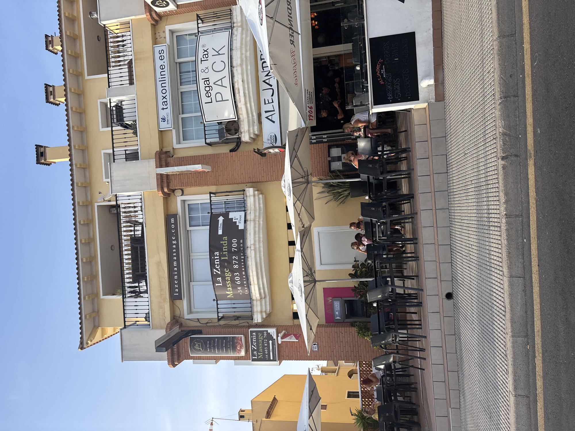 The image shows a street-level view of a yellow building with balconies and awnings in Urbanización La Cenia II, Spain. The building houses several businesses, including a massage salon with signage for "La Zenia Massage - Linda" and a website address "lazzeniamassage.com". Next to it is a sign for "Legal & Tax PACK" and "taxonline.es". A restaurant named "ALEJANDRO'S BAR RESTAURANTE" is visible at street level with outdoor seating. Several people are seated at tables outside the restaurant under large white umbrellas, some appearing to be dining or socializing. An ATM machine is also visible. The building has a light blue sky above and a sidewalk and street in the foreground.