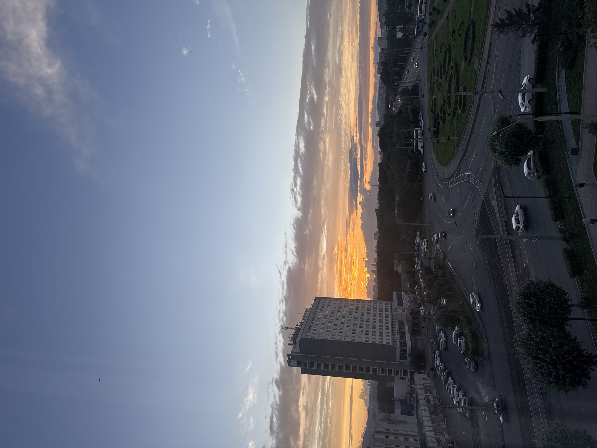 This image shows an aerial view of a city intersection in Krasnoyarsk, Russia, during a sunset. A tall, modern building, possibly a hotel or office building, is prominently featured on the left side of the frame. It is a grey, multi-story structure with many windows and an antenna on its roof. Surrounding the building are other urban structures, including lower-rise buildings and a parking lot filled with cars.
The intersection itself is a complex network of roads with multiple lanes, traffic lights, and streetlights. Several cars are visible on the roads, some in motion and some parked. There are also pedestrian walkways and green spaces, including a circular area with manicured bushes. The sky is a gradient of blue and orange, with scattered clouds illuminated by the setting sun, creating a dramatic and colorful backdrop. In the distance, a cityscape with more buildings is visible, extending towards a tree-covered horizon. The overall scene depicts a busy urban environment at dusk, with a focus on the intersection and the surrounding architecture.