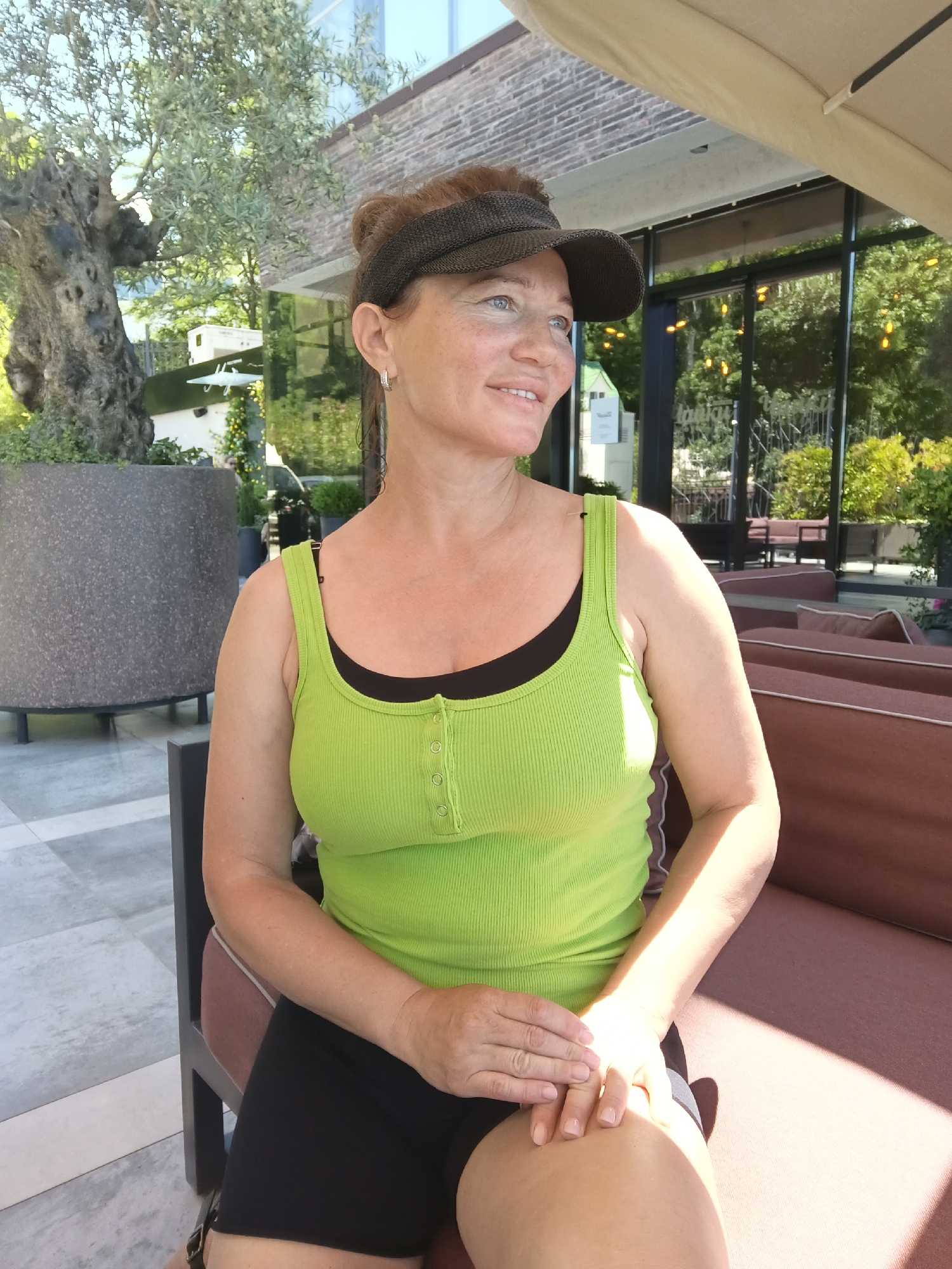 A middle-aged Caucasian woman sits outdoors on a cushioned bench, her body angled slightly to her right. She wears a lime green ribbed tank top over a black bra and black shorts, with a brown visor on her head. Her hair is pulled up in a bun. Her left arm rests on her lap, with her hands crossed. Her gaze is directed to her right, and she has a slight smile. The background shows a large potted tree to the left, a brick building with large windows, and outdoor seating with brown cushions to the right. Sunlight streams in from the upper right, casting shadows on the woman and the bench. The environment suggests a restaurant or lounge area in an urban setting.