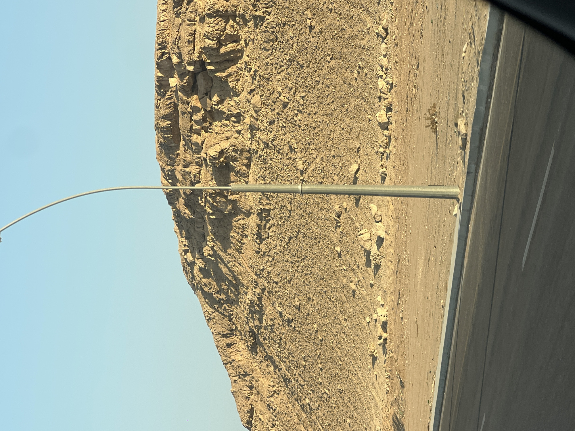 The image depicts a desert landscape in Sharm El Sheikh, Egypt, featuring a rugged, rocky hill in the background under a clear blue sky. A solitary streetlight stands prominently in the foreground, positioned near the edge of a paved road. The road itself is dark asphalt with white lane markings. The ground surrounding the hill and the road is sandy and arid, dotted with scattered rocks and a small bush. The lighting suggests it is daytime, likely late afternoon or early morning given the long shadows cast by the streetlight.