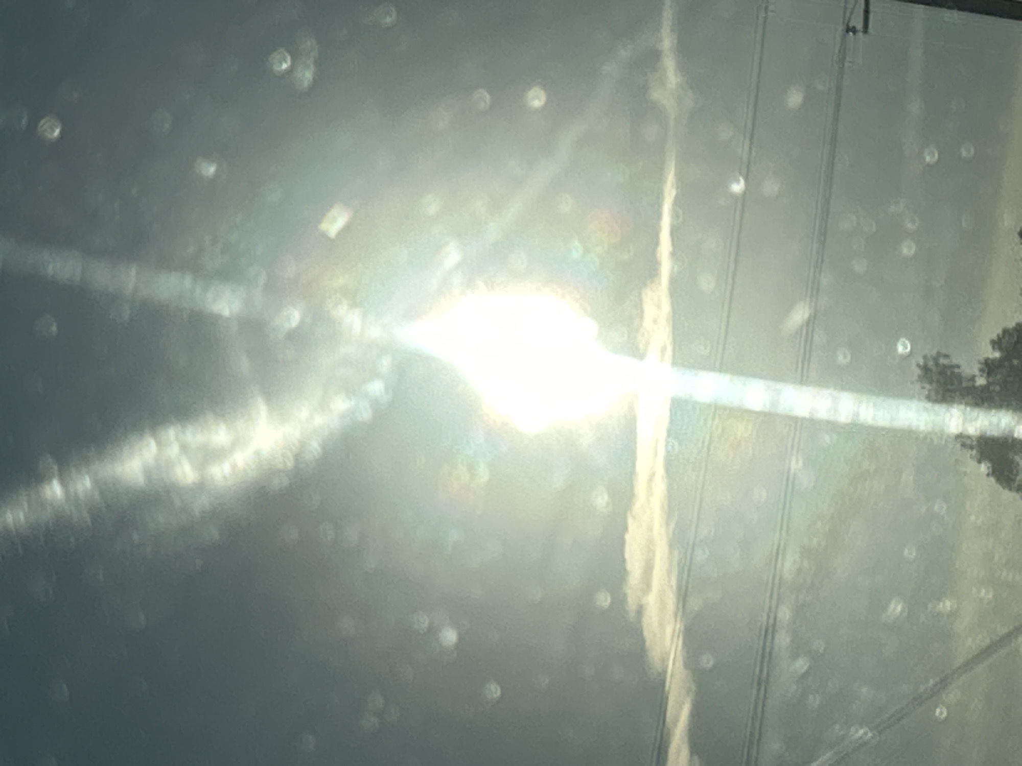 The image displays a bright, hazy sun obscured by what appears to be a dirty or streaked window, possibly from a vehicle. The sun is the most prominent element, creating a lens flare effect with a vertical streak of light extending upwards from it. Numerous circular or oval light refractions, likely due to water droplets or dust on the window, are scattered across the frame. In the middle ground, a thin band of clouds is visible, with horizontal lines that could be power lines. The lower portion of the image shows a blurry outline of trees and a utility pole. The overall scene suggests an outdoor environment with overcast or hazy conditions, viewed through a compromised surface. There are no people or clear actions depicted. The location in Matrayevo, Russia, is not discernible from the visual information alone.