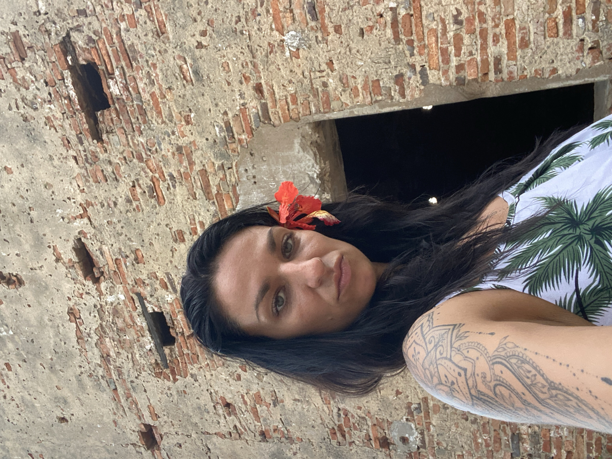 The image shows a woman with long dark hair, wearing a floral shirt and a red flower in her hair. She is positioned in front of a weathered brick wall with several dark openings. The location is Phu Thuy Ward, Vietnam, as indicated by information provided with the image. She appears to be in an old, possibly abandoned structure, given the condition of the wall. The woman has tattoos on her arm.