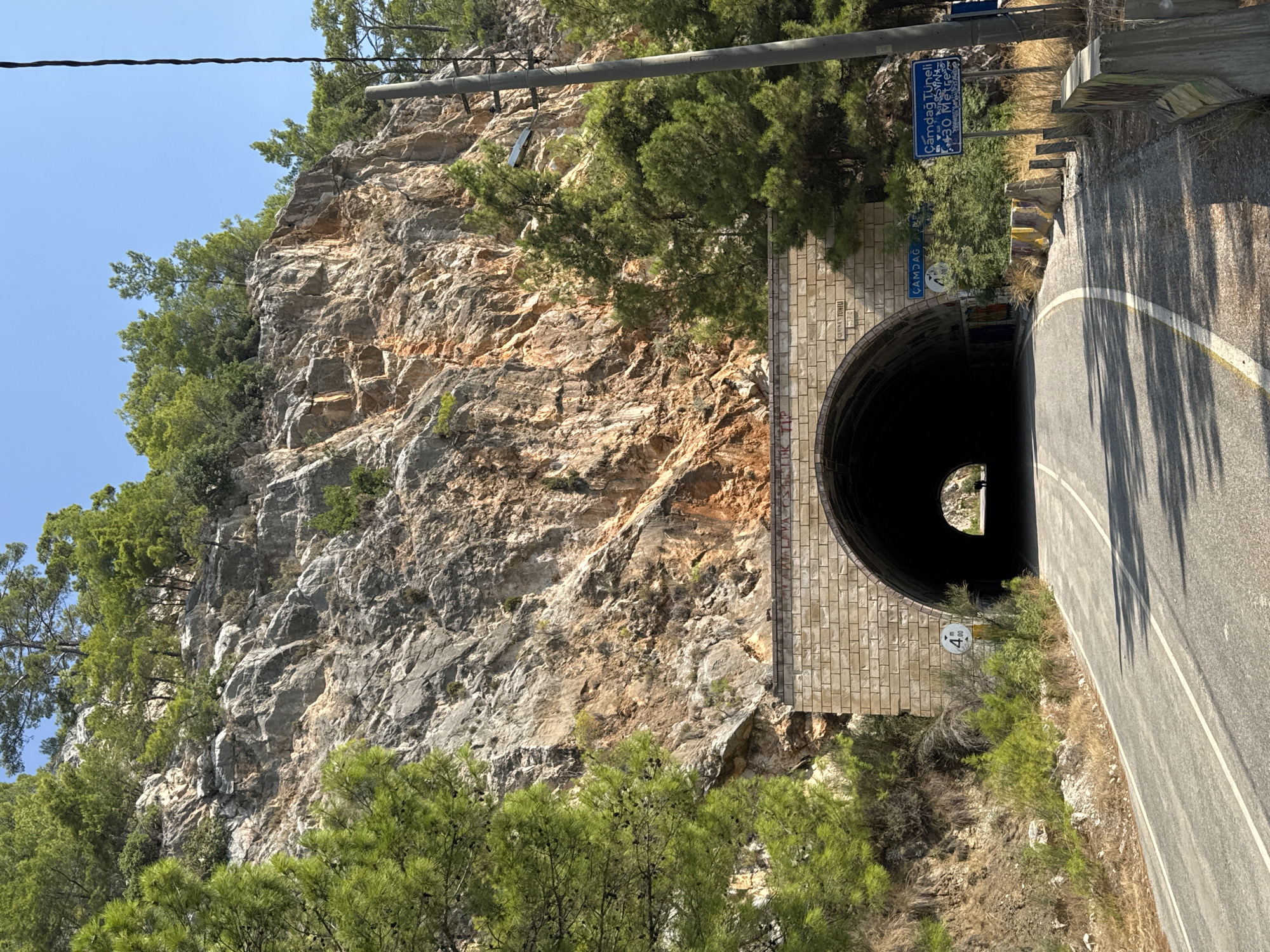 The image depicts the Çamdağ Tunnel in Kemer, Turkey, a road tunnel carved into a rocky mountainside. The tunnel entrance is constructed of light-colored stone blocks, forming an archway. A sign to the left of the entrance indicates a height limit of 4.00 meters. To the right of the entrance, blue road signs are visible, one with Turkish text that reads "Çamdağ Tüneli" and "30 Metre", indicating the tunnel name and length. The road leading to the tunnel is paved and curves slightly. The surrounding environment is mountainous with sparse vegetation, including pine trees and shrubs. The sky is clear and blue. Two figures are visible walking within the dark interior of the tunnel, appearing as silhouettes against the light at the far end. There are also some graffiti markings on the tunnel entrance. The overall scene captures a section of road infrastructure in a natural, mountainous landscape.