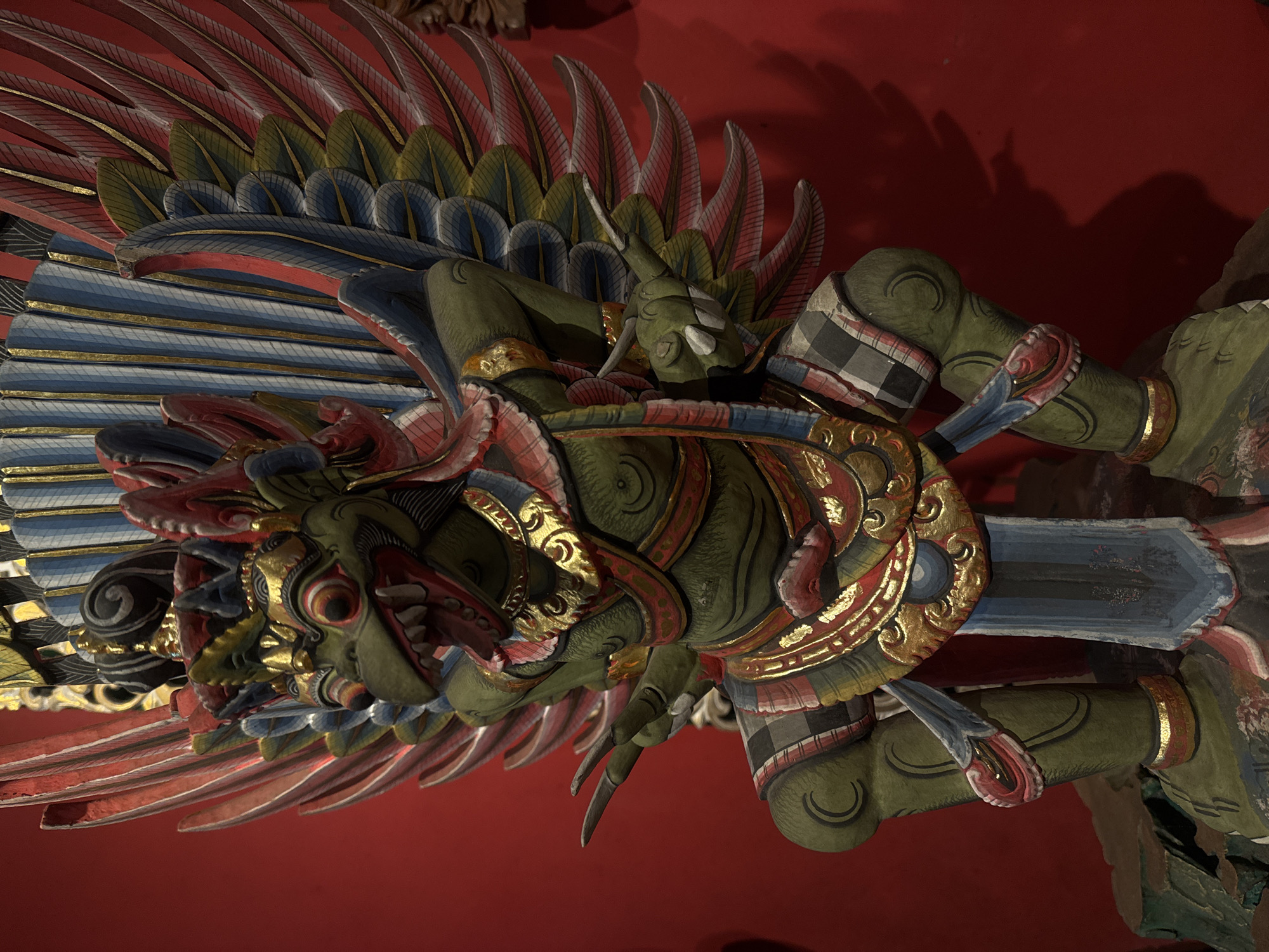 This media file features a close-up, angled view of an intricately carved and painted wooden sculpture, likely a Garuda or a similar mythical bird-like creature from Balinese art. The sculpture is predominantly green, with accents of red, gold, blue, and black. Its wings are spread wide, composed of numerous layers of feathers rendered in vibrant colors. The creature's face is detailed with bulging eyes, sharp teeth, and a prominent beak. It is adorned with ornate jewelry, including gold bands around its arms and legs, and decorative chest armor. The sculpture is suspended against a rich red background, possibly a ceiling or wall, which is out of focus. The lighting appears to be artificial, casting subtle shadows and highlighting the textures and details of the artwork. The overall impression is one of grandeur and traditional artistry. There is no visible text in the image. Given the location context of Kuta, Indonesia, this artwork is likely found in a temple, a cultural performance venue, or a decorative architectural element in a building.