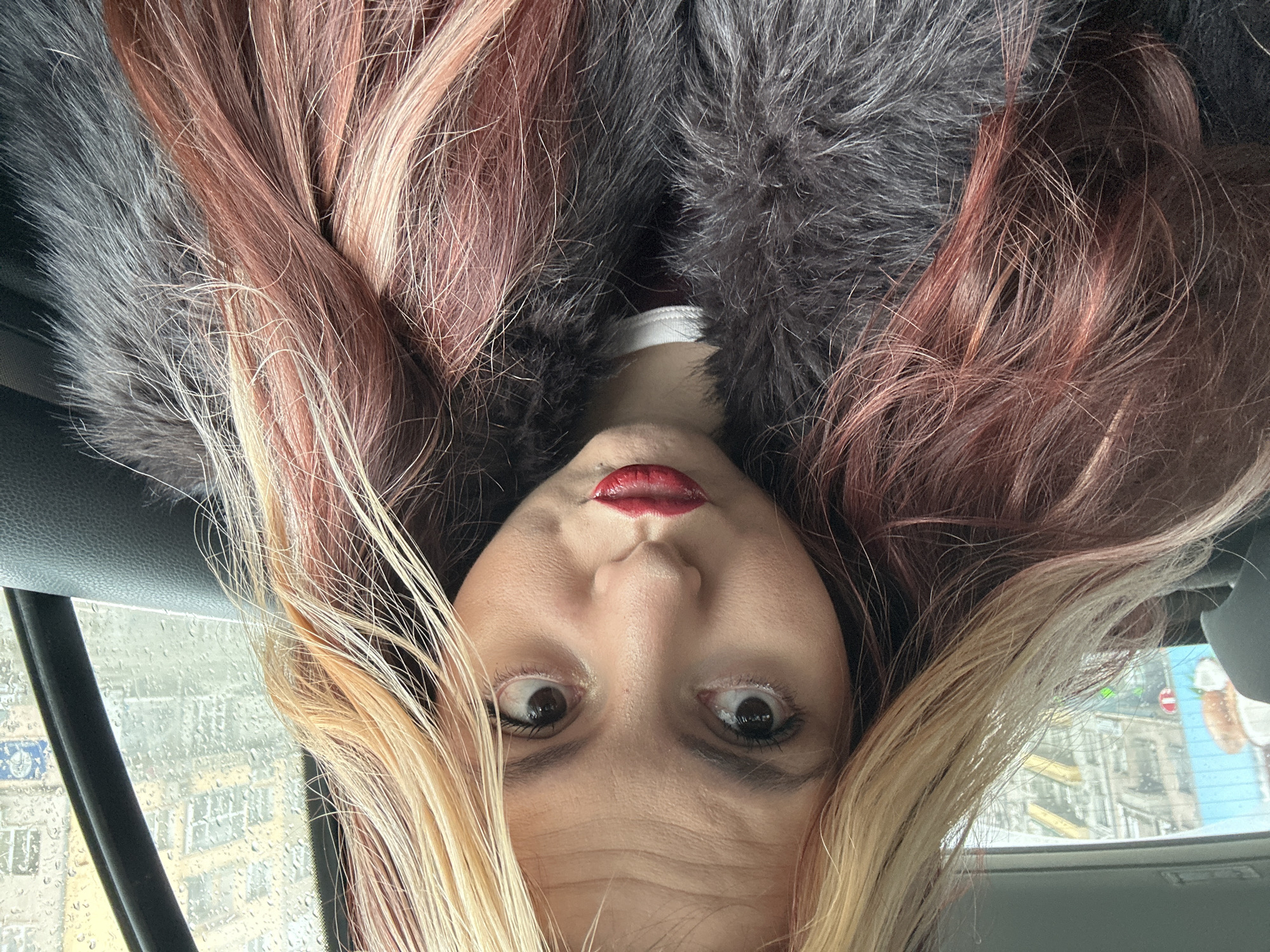 A selfie photograph of a woman with red lipstick, taken from an inverted angle, possibly inside a vehicle. The background shows a rain-streaked window with an obscured view of a city street. The woman is surrounded by dark gray fur and blonde and red hair. The city is possibly Sofia, Bulgaria, but this cannot be confirmed from the image.