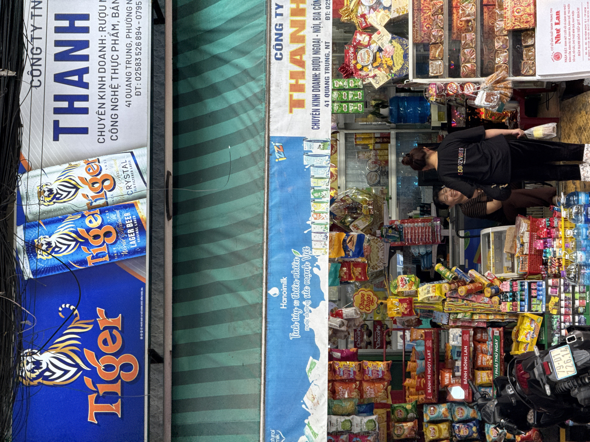 A street-level view captures a storefront in Nha Trang, Vietnam, displaying an array of products. Above the shop, a large blue and white banner advertises "Tiger" lager beer with imagery of tigers and beer cans. To the right, another banner for "THÀNH" and "CÔNG TY TN" indicates a business selling foreign liquors, food products, and other items. The storefront itself is packed with merchandise, including bags of chips, snacks, drinks, and gift baskets. A dark-colored motorcycle is parked in front of the shop. Inside the store, two women are visible: one, wearing a black t-shirt and pants, appears to be a shop employee holding a plastic bag, while the other, seated and wearing a striped shirt, is smiling. The environment suggests a bustling commercial area with a mix of local and imported goods.
