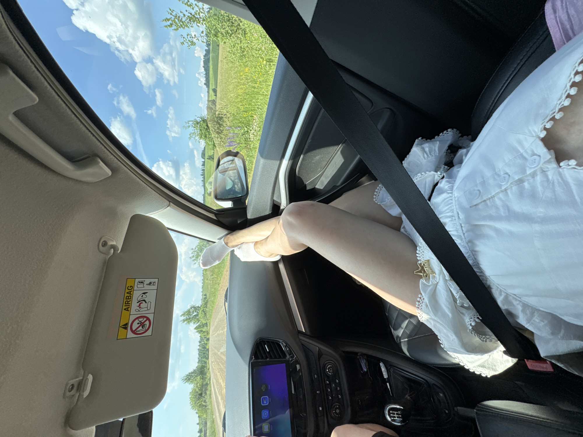 The image shows the interior of a car with a person's legs propped up on the dashboard, visible through the open passenger window. The exterior scenery suggests a rural or semi-rural environment with fields and trees under a bright, cloudy sky. The car's interior includes a sun visor with an airbag warning sticker, a dashboard with a central infotainment screen and controls, a gear shift lever, and a seatbelt. The person is wearing white socks and a white garment with lace trim, adorned with a small star-shaped clip. The car appears to be on a dirt road, consistent with a location outside a major urban center. The city of Van'ki, Russia, is a village, so this setting aligns with the visual information.