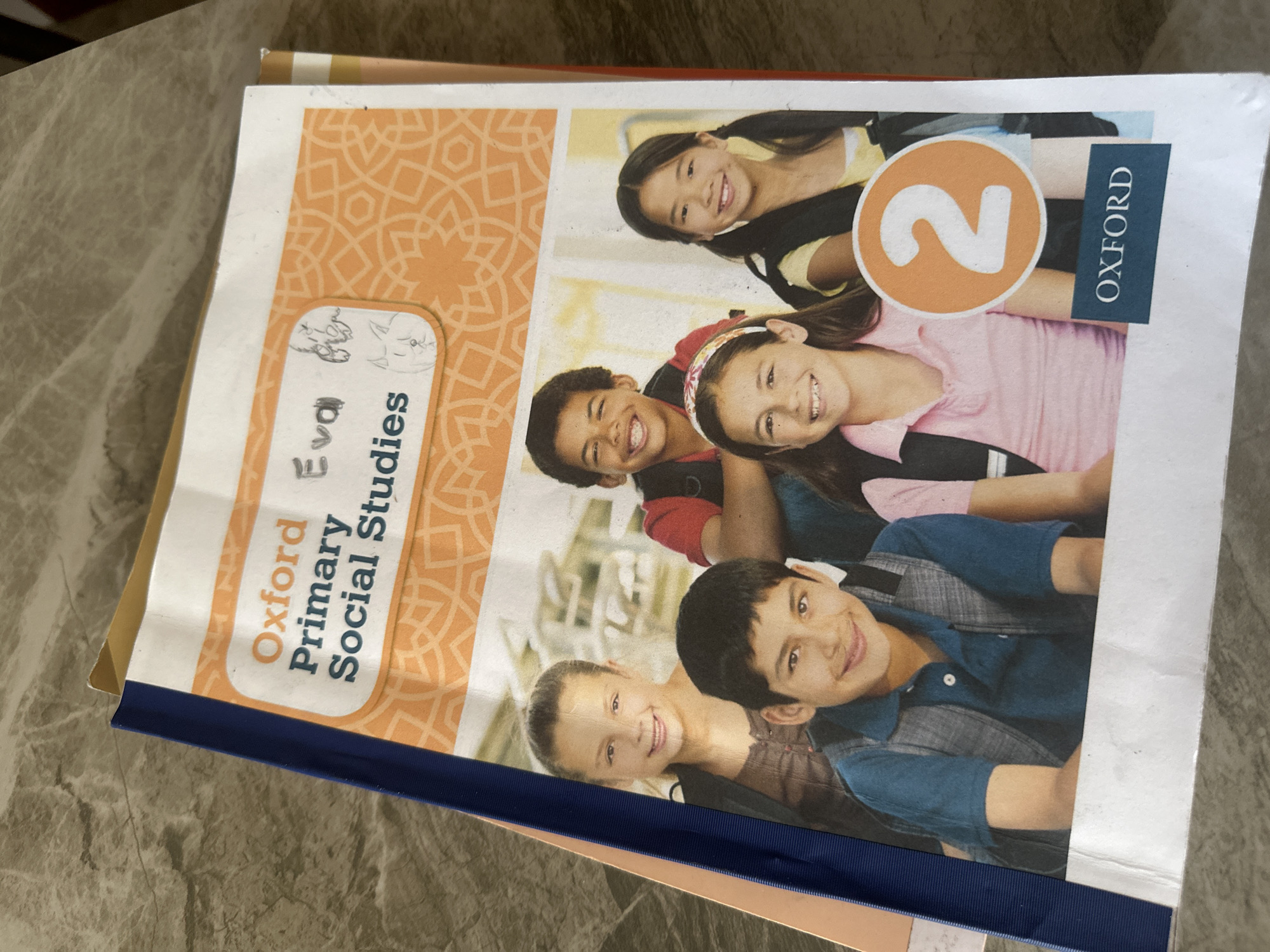 The image shows a textbook titled "Oxford Primary Social Studies 2." The textbook cover features a patterned orange background on the left side. The right side of the cover has a photograph of five smiling children of different ethnicities. The number "2" is prominently displayed in a large orange circle, and the word "OXFORD" is printed in a vertical blue box. A name, "Eva," is written in marker on a white oval label on the upper-left area of the cover. The textbook has a dark blue binding and is placed on a light-colored surface. There is no information in the image to indicate the location is Bac Nha Trang Ward, Vietnam.