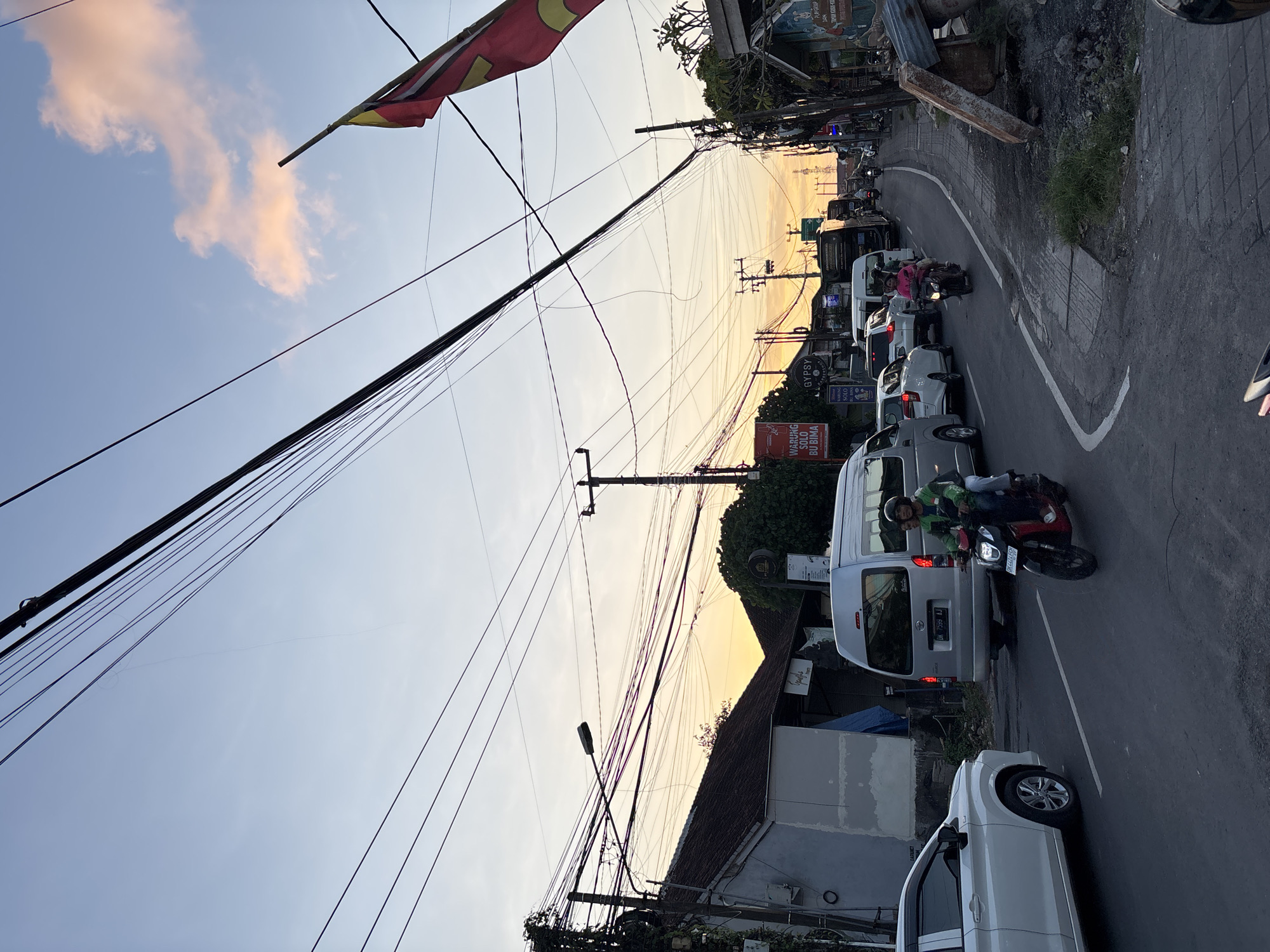 The image depicts a street scene in Kuta Selatan, Indonesia. Traffic is congested with a line of cars and motorcycles. A person wearing a green shirt rides a motorcycle in the foreground. A flag hangs overhead in the upper left corner. Power lines stretch across the sky. The sky is light with a hint of sunset. Buildings and vegetation border the road.