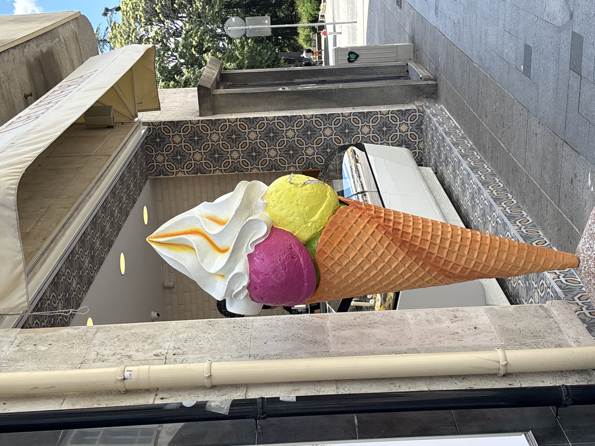 An oversized, realistic sculpture of a triple-scoop ice cream cone stands prominently outside an establishment. The cone itself is a textured, orange waffle cone. The scoops are a vibrant fuchsia, lime green, and topped with swirled white soft-serve ice cream drizzled with orange syrup. The sculpture is situated on a sidewalk with decorative tiled borders. Behind the ice cream cone, the building features a grey stone facade and a patterned tile accent wall. An awning, partially visible at the top, is a cream color. To the right of the building entrance, a street scene unfolds with trees and a few individuals walking in the distance. The context provided suggests the location is Burgas, Bulgaria.