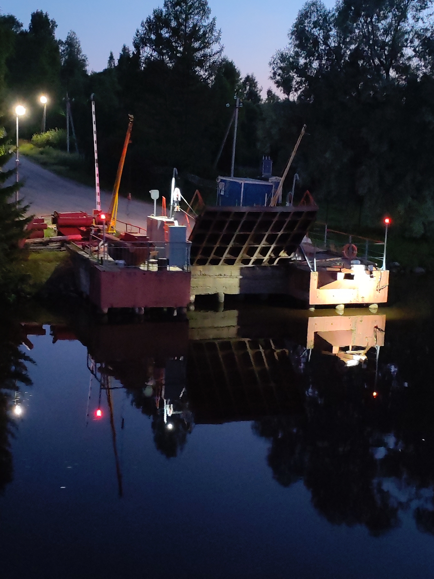At dusk in Annensky Most, Russia, a ferryboat with a red ramp is docked. The ramp is partially raised, showing a diamond-shaped grid pattern. The ferryboat has a red structure with an orange crane extending from the left side, and a blue cabin on the right. Lights are visible on the ferryboat, and a red signal light is on a pole on the right side. A red life preserver is attached to a railing. The ferryboat is reflected in the dark water. On the shore, a road is visible with streetlights and a white and red barrier. Trees and foliage surround the scene, with a dark blue sky overhead.