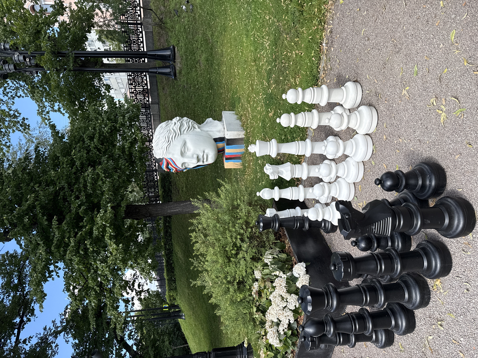 The image depicts a large outdoor chess set situated in a park-like environment. The chess pieces are oversized, with both black and white sets visible in the foreground. Behind the chess set, a statue of a human head, appearing to be made of white stone, is decorated with colorful stripes on its forehead and base. Lush green trees and grass surround the statue and chess set, suggesting a summer or early autumn setting. In the background, a black decorative fence runs along a raised embankment, and buildings can be seen beyond the trees. The overall scene appears to be in a public urban space, likely a park or square. The presence of a statue and large-scale art installation indicates a location that values public art and recreation. Based on the artistic style of the statue and the overall aesthetic, the location is likely in Moscow, Russia, potentially in a well-known cultural or recreational area. There are no people visible in the immediate foreground of the image, but some pigeons are visible on the grass in the background.