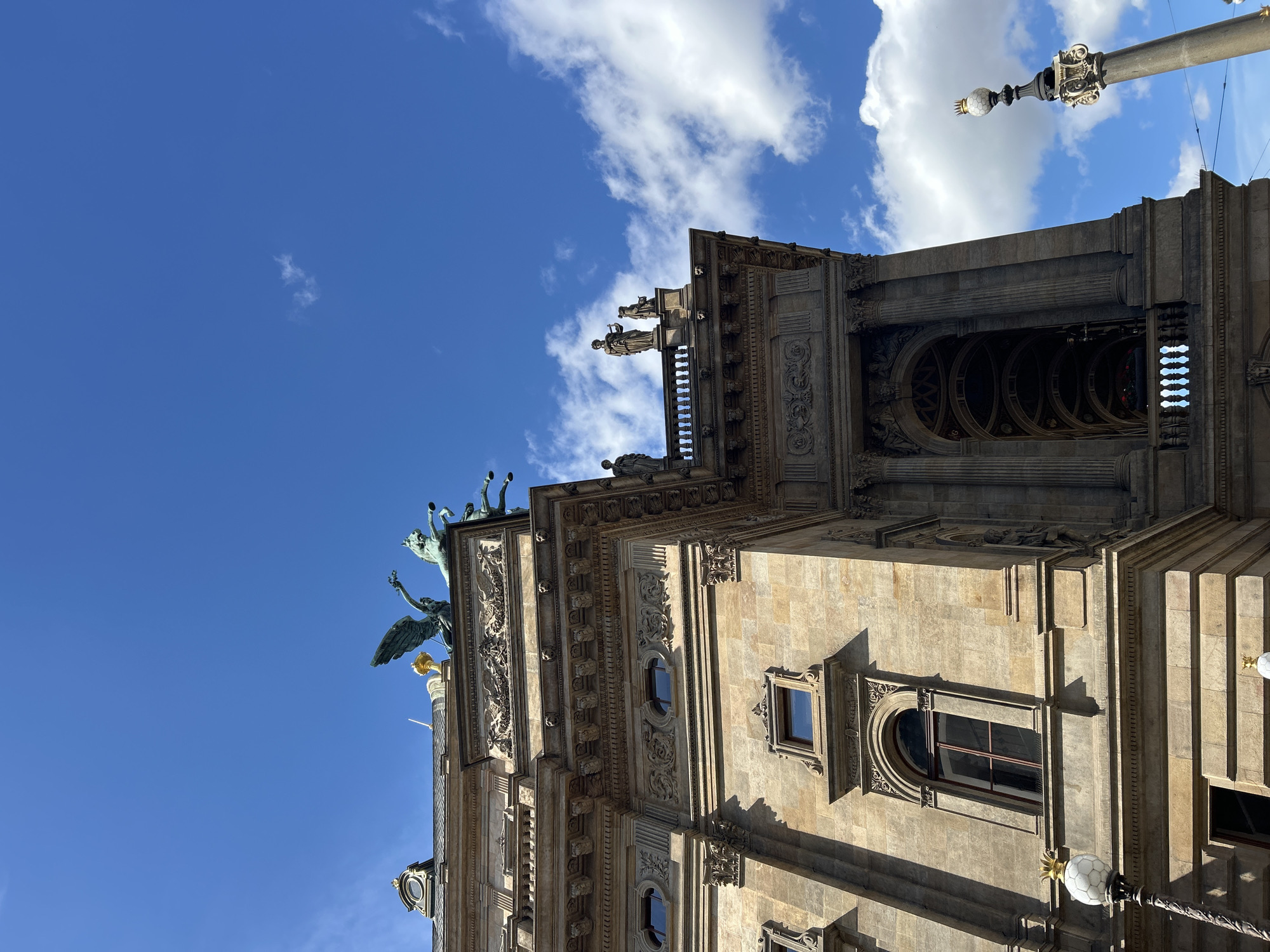 An upward angle shot captures the ornate facade of the Estates Theatre in Prague, Czechia, under a bright blue sky dotted with fluffy white clouds. The theatre's sandstone exterior is adorned with intricate carvings and classical architectural elements. Atop the building, a bronze sculpture depicts a chariot pulled by horses, with winged figures, and statues of individuals are visible on a balcony. A lamppost with a decorative globe stands to the right of the building. The image is taken on a sunny day, with clear visibility of the architectural details and the sky.