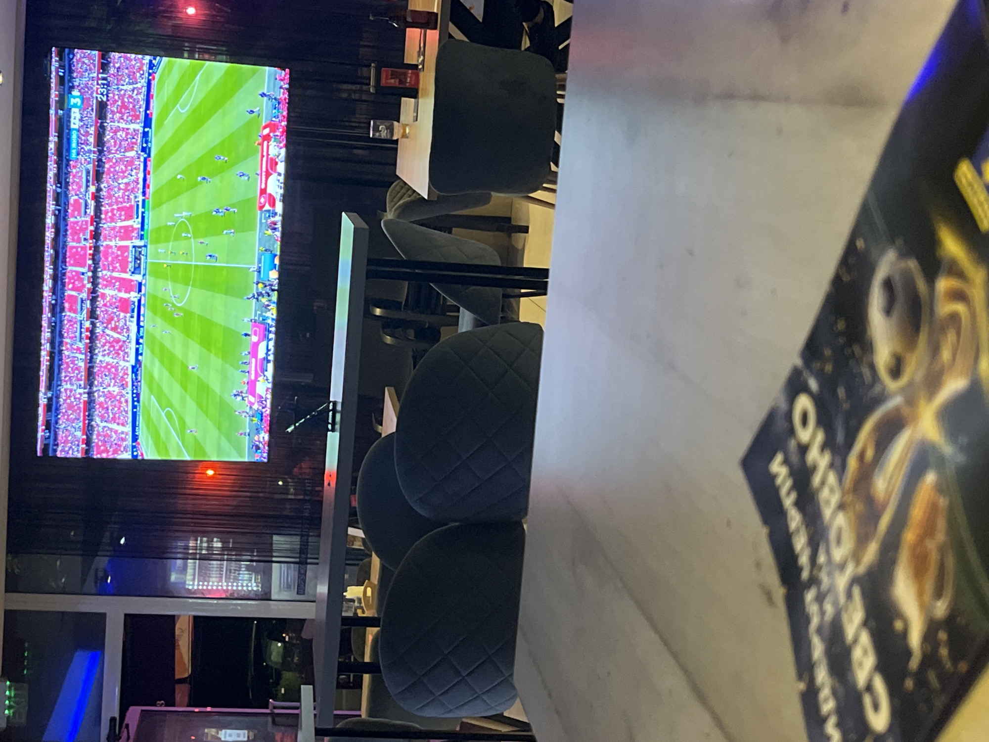 An interior shot captures a stadium screen displaying a soccer game in a dimly lit bar or lounge. The screen shows a wide-angle view of a soccer field with players and spectators. In the foreground, a table is partially visible with a book on it, featuring text that appears to be in Cyrillic. Several dark upholstered chairs are arranged around tables, some of which are made of dark wood. A glass of beer and a bottle are on one of the tables. The environment suggests a casual social setting where patrons can watch sports.