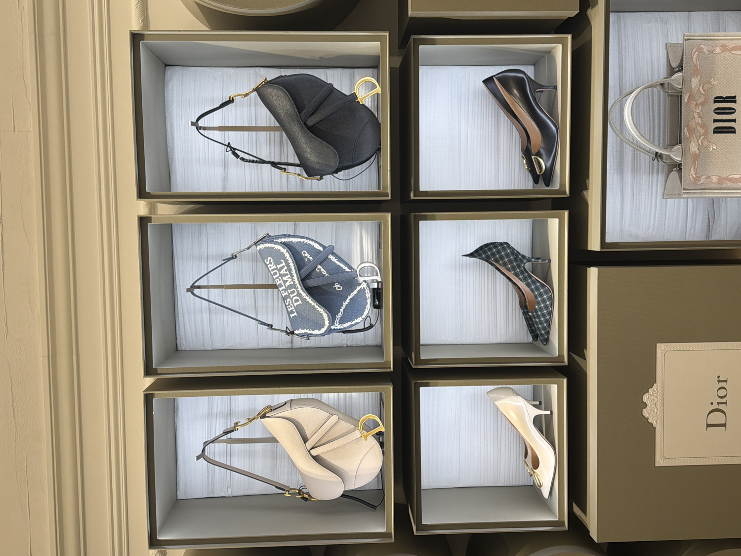 This image captures a refined retail display showcasing an assortment of luxury Dior handbags and high-heeled shoes, indicative of a high-end boutique setting, possibly in London, UK. The items are meticulously arranged within individual, uniformly lit display boxes, which are framed by sleek, darker-toned surrounds.
On the left, three iconic Dior Saddle bags are displayed vertically. The top bag is black with gold hardware. Below it is a blue denim-style Saddle bag, embroidered with the text "LES FLEURS DU MAL" and a stylized "Dior" logo, also featuring gold hardware. The bottom Saddle bag is a light beige or off-white color, completed with matching gold hardware.
The right side of the display features three pairs of elegant Dior pumps. The top pair consists of black high heels with pointed toes and a gold buckle, likely bearing the 'CD' emblem. Beneath them, a pair of plaid high-heeled pumps, possibly in green and black, are artfully positioned. The bottom pair are cream or off-white high heels, similar in design to the black ones with a distinct gold buckle.
Partially visible on the far right, above and below the shoes, are other Dior products. A light-colored Dior Book Tote bag, embroidered with "DIOR" and a decorative floral motif, is seen at the top right. Below the plaid shoes, a vertical "Dior" logo is present on a white label, set against a larger, dark box. The backdrop includes a light beige, vertically fluted wall or column on the far left. The scene is static, devoid of people, and highlights the luxurious aesthetic of the brand's offerings.
