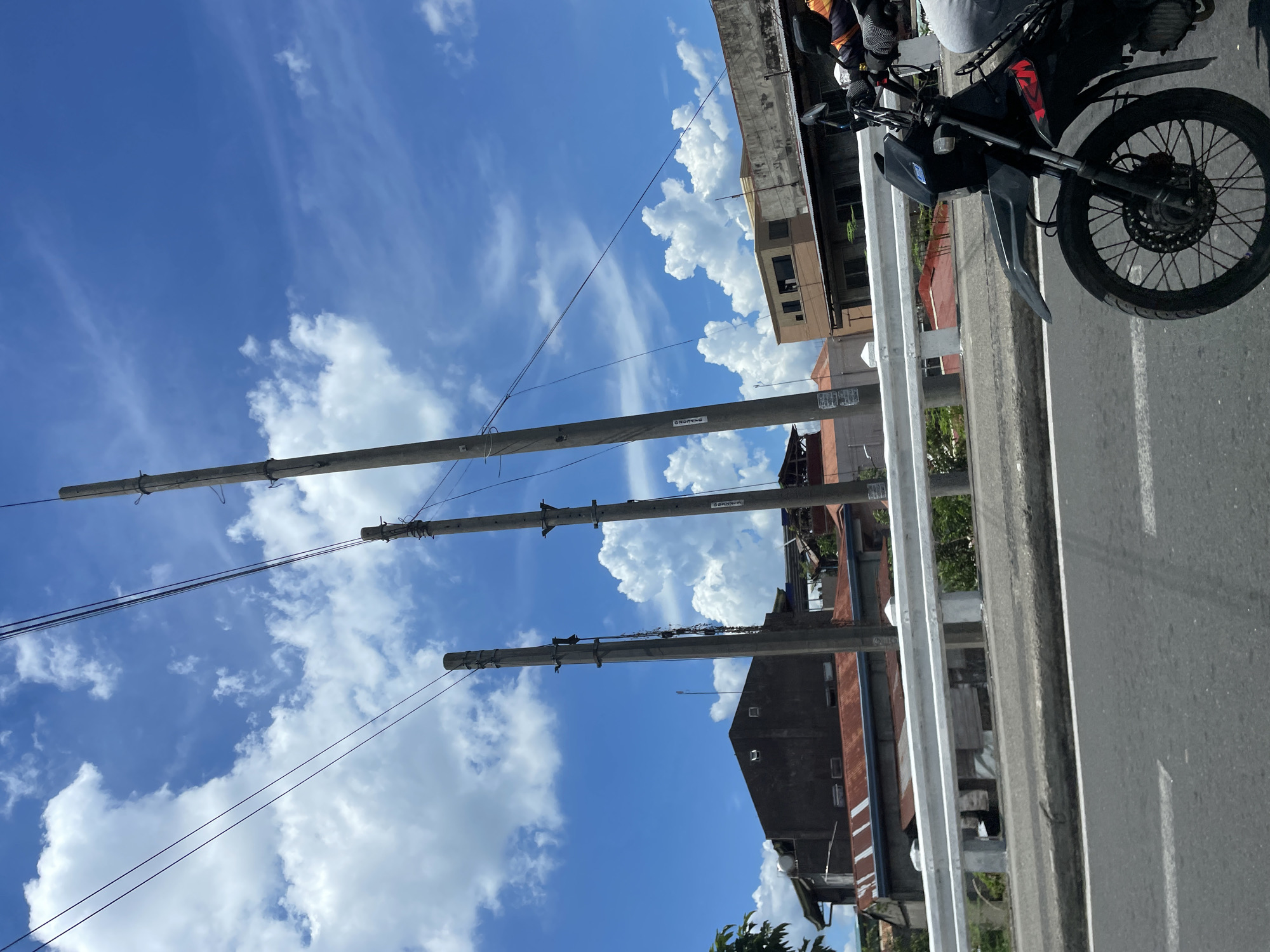 A motorcycle is moving on a road in Davao City, Philippines, with a blue sky and white clouds overhead. The road is paved and has white lane markings. A white metal barrier runs along the side of the road. Beyond the barrier, there are several buildings with corrugated metal roofs, some with a reddish-brown color. Utility poles with wires are visible, some with labels attached. The motorcycle is black with red accents and is partially visible, showing its front wheel, handlebars, and a rider's arm and leg. The rider is wearing a jacket with colorful stripes. The scene suggests a bright, sunny day in an urban or semi-urban area.