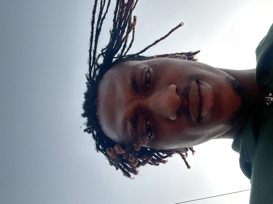 An individual with dark skin and dreadlocks is captured in a low-angle, close-up portrait in Ilishan, Nigeria. The subject, appearing male, looks directly into the camera with a neutral expression. Dreadlocks are visible on the head; some on the left side are shorter and clustered, while those on the right are longer and appear to be swept towards the right, suggesting environmental airflow. Minimal stubble is present on the chin and upper lip. The subject wears a dark green hooded garment, revealing a silver-colored zipper pull at the collar. The background is a bright, largely clear sky, transitioning from a lighter glow on the lower left to a faint blue-grey towards the top. A thin dark line, possibly a wire, is visible vertically along the far left edge of the frame.