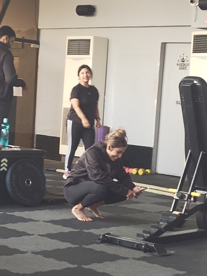 This image captures a vibrant scene inside a fitness studio or gym, likely in Kolkata, India, during daytime. The tiled dark grey floor and white walls suggest a modern and clean environment.
Three individuals are present. In the immediate foreground, a woman with light brown hair, styled in a bun, is barefoot and in a deep squat position. She is dressed in a black hoodie and black leggings, holding a smartphone and smiling warmly as she looks at the screen. To her right, part of a black and yellow exercise machine is visible. To her left, several heavy black weight plates are stacked against a black plyometric box, and a blue water bottle rests on a surface.
Standing behind the squatting woman, towards the center of the frame, is another woman who is also smiling, looking in the general direction of the first woman or the camera. She wears a black t-shirt and black track pants with white stripes down the side and appears to be holding a rolled-up purple yoga mat or towel in her right hand.
On the far left, partially visible, is a man in a dark top, looking down at some papers or notes he is holding.
The background features white walls with two prominent white standing air conditioning units. A white door on the right side of the image displays a sign featuring a lotus-like symbol above the text "MASSAGE ROOM." Below this, the letters "SL" are partially visible. Further back on the floor, some colorful smaller weights or resistance bands can be seen. The overall atmosphere appears casual and friendly, with individuals engaged in their activities.