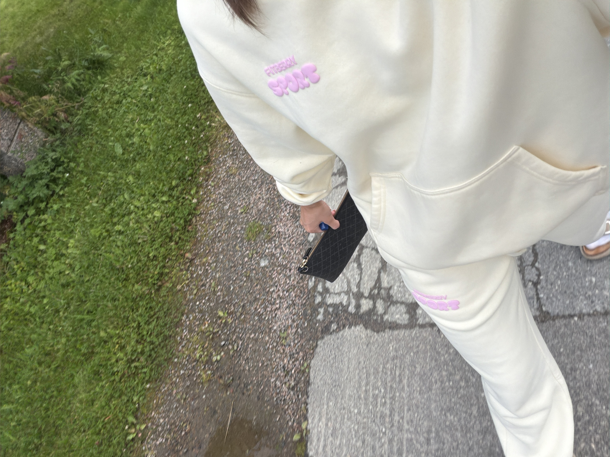 A person wearing a cream-colored tracksuit with pink lettering on the left leg and back of the sweatshirt is walking on a sidewalk next to a patch of grass. The person is holding a black quilted clutch in their right hand. The ground is wet in places, suggesting recent rain. The scene is outdoors in what appears to be a residential or park area. The location is not specified to be in Harstad, Norway, as there are no visible landmarks or contextual clues to support this.