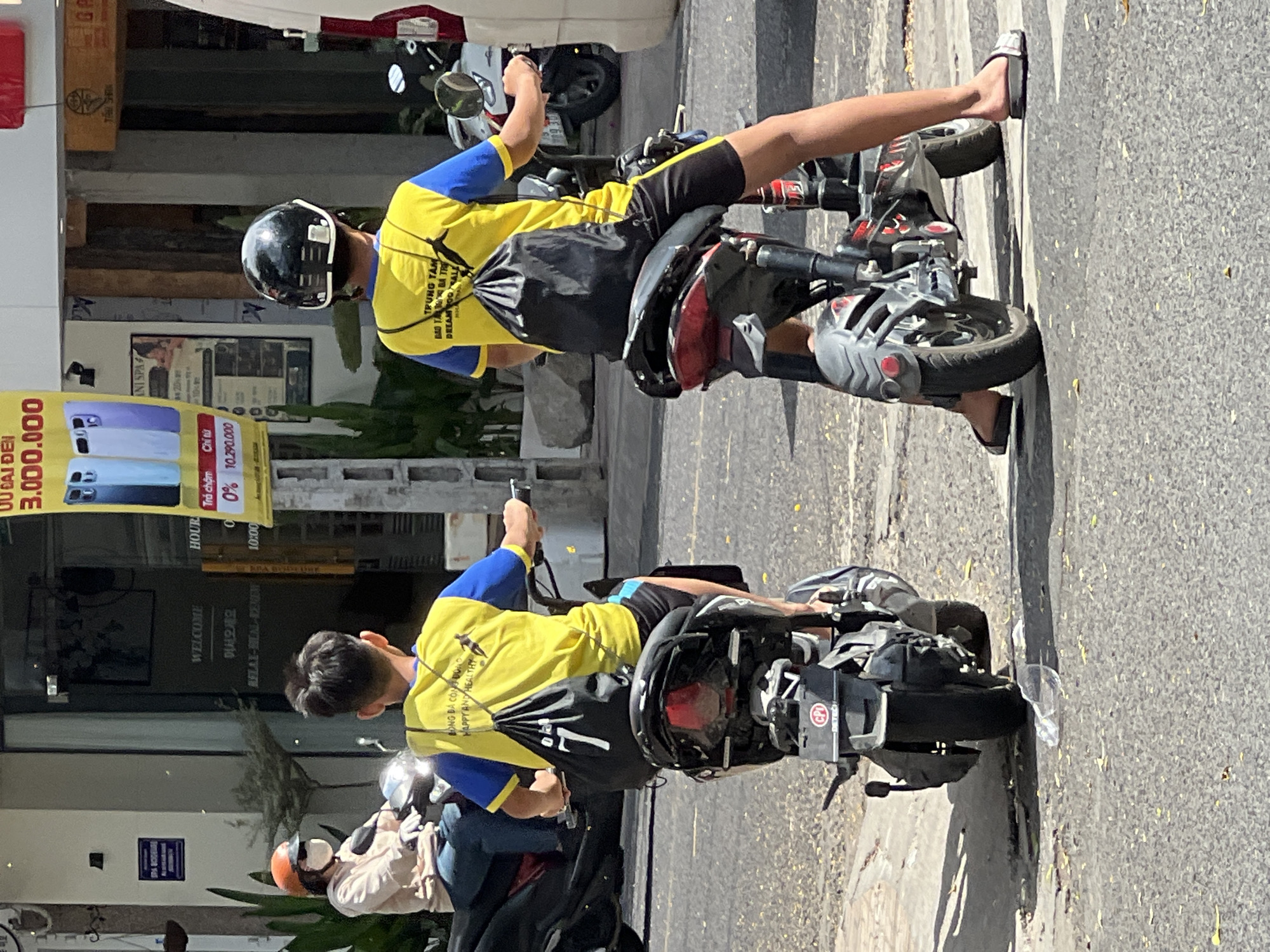 Two individuals on electric scooters are seen on a street in Nha Trang, Vietnam. The person on the left, wearing an orange helmet, a light-colored hooded sweatshirt, and blue jeans, is riding a black scooter. The person on the right, wearing a black helmet and a yellow and blue shirt with black shorts, is riding a black electric scooter with red accents. This individual has a black drawstring bag with the number "7" on it hanging over their shoulder and a black backpack on their back. The background shows buildings with signage, including "WELCOME" and advertisements. The street is paved, and there are some plants visible. The lighting suggests it is daytime.
