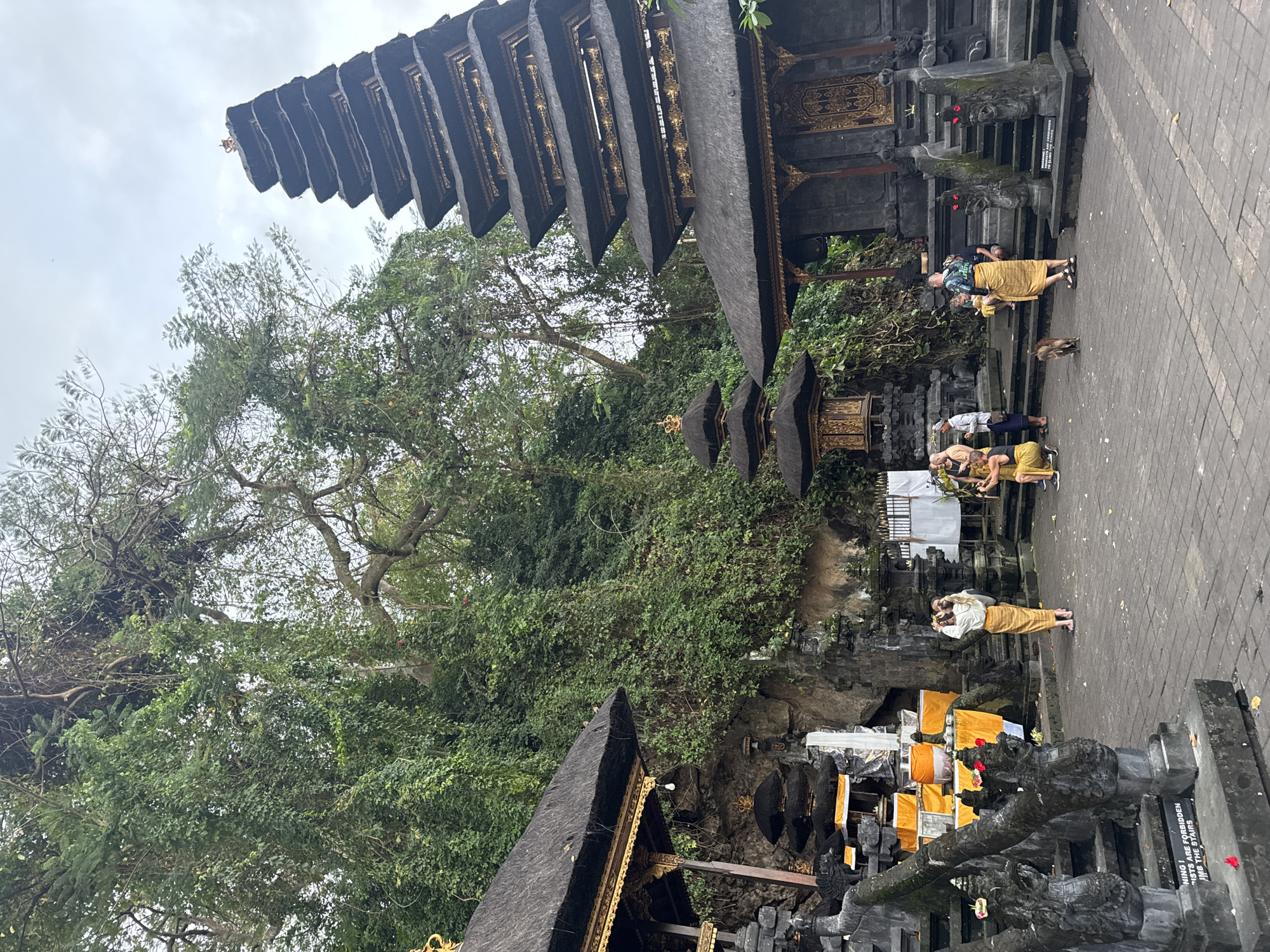 In the city of Dawan, Indonesia, an outdoor scene unfolds at a temple complex. The architecture features tiered, dark stone structures with thatched roofs, intricately decorated with gold accents. A prominent multi-tiered pagoda-like building dominates the right side of the frame, with a smaller, similar structure to its left. In the background, a lush, verdant hillside is covered in dense foliage and trees, partially obscuring the sky.
Several individuals are present in the courtyard. On the left, a woman in traditional Balinese attire, a cream-colored top and a yellow sarong, stands with her back to the camera, holding a phone and appearing to take a photo. In the center, two figures in similar yellow and cream traditional clothing are interacting, one appearing to be seated. To the right, a person in a dark shirt and a yellow sarong is observed. A small, light brown cat is in the foreground, walking away from the camera. The ground is paved with grey interlocking stones, and stone carvings of mythological creatures line a stairway on the left. Several offerings wrapped in yellow fabric are visible on pedestals. A sign with red text is partially visible at the base of the stairs on the left. The overall atmosphere suggests a cultural or religious site.