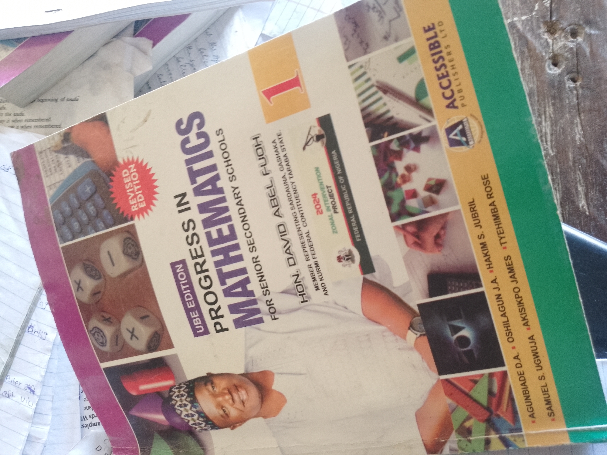 The image is a close-up, angled, exterior shot of a book titled "Progress in Mathematics for Senior Secondary Schools". The book is a revised edition and features the number "1" prominently on its cover, along with an image of a man's face and various educational illustrations. The book is resting on a stack of other papers and books, some of which have handwritten notes. The environment appears to be indoors, possibly a classroom or study area, with a wooden table visible in the background. The specific location in the city of Serti, Nigeria, is not discernible from the image itself.