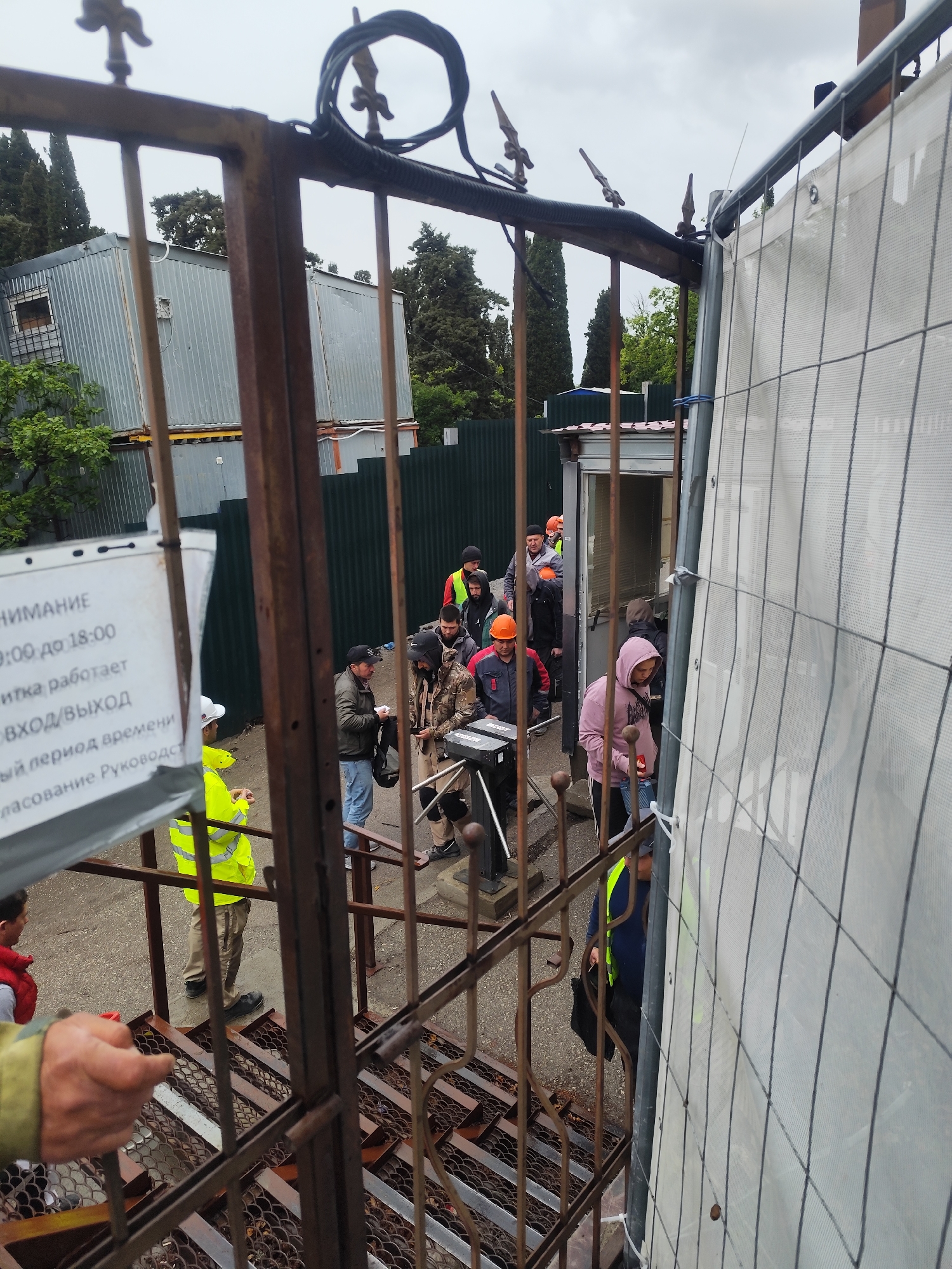 The image captures an outdoor scene at what appears to be the entrance or exit of a secured work area, possibly a construction site or industrial facility, in Симеизский поселковый совет, Ukraine. The photograph is taken from a slightly elevated perspective, looking through a rusty, ornate metal gate in the foreground.
Numerous individuals, predominantly men, are visible near a turnstile and a small security booth, suggesting a controlled access point. Many are dressed in work attire, including hard hats (orange, white) and high-visibility vests (yellow, orange), indicating they are workers. One person in a bright yellow vest and white hard hat stands to the left, while another in a pink hoodie is closer to the turnstile. People appear to be queuing or passing through the access point in a routine manner.
To the right, a temporary chain-link fence covered with a white mesh fabric partially obscures the view. In the background, several grey corrugated metal containers are stacked, likely serving as temporary offices or storage. Tall, slender evergreen trees are visible under an overcast sky, indicating daylight hours.
A white sign attached to the metal gate displays Russian text. Visible portions include:
* "ВНИМАНИЕ" (ATTENTION)
* "9:00 до 18:00" (9:00 to 18:00)
* "ВХОД/ВЫХОД" (ENTRY/EXIT)
* Partial text like "...тика работает" (turnstile operates/pass works), "...ый период времени" (other period of time), and "...сование Руководст" (agreement with Management), likely detailing access rules and operating hours.
The scene depicts a typical workday entry/exit point, with a focus on access control for personnel.