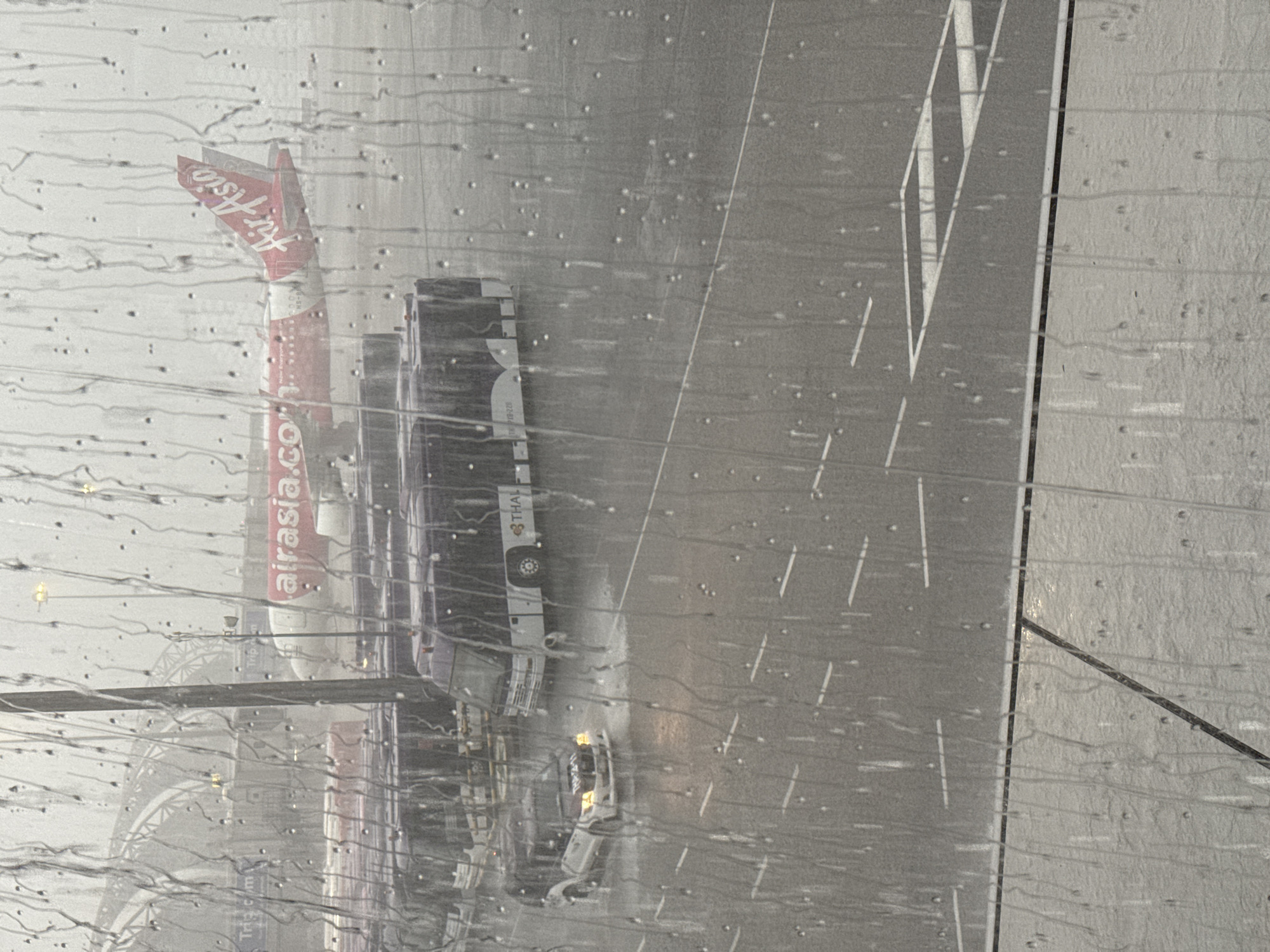 This image shows an airplane viewed through a window covered in raindrops. The airplane is red and white, with "AirAsia.com" printed on its side. Behind the AirAsia plane, several white buses with "THAI" written on them are parked. The scene appears to be an airport tarmac in Nong Prue Subdistrict, Thailand. The weather is rainy, indicated by the water droplets on the window and the wet appearance of the tarmac. The lighting suggests it is daytime, possibly overcast due to the rain. The overall mood is somewhat subdued, likely due to the weather.