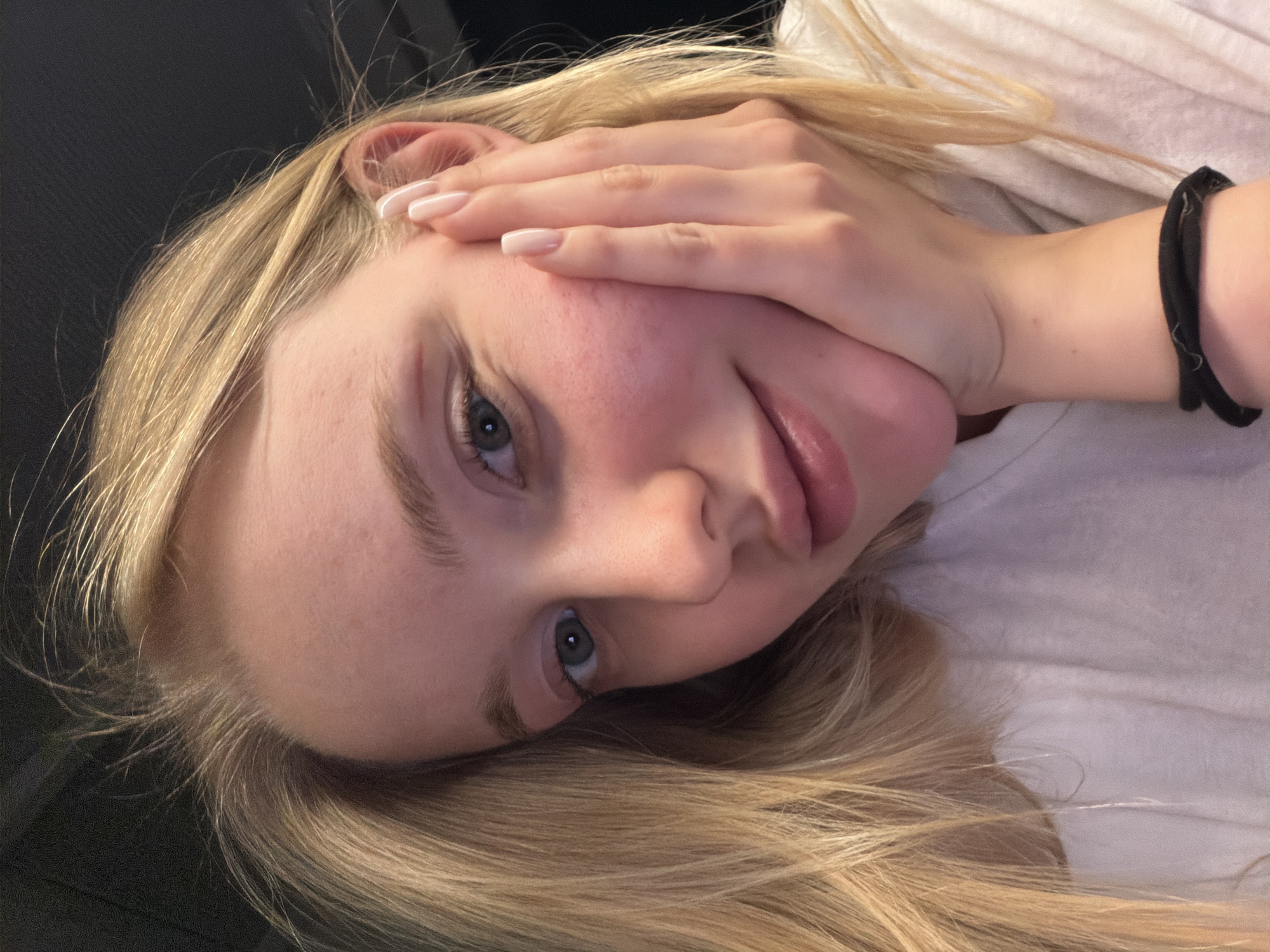 A close-up portrait shows a young woman with light blonde hair, light skin, and blue eyes. Her head is tilted to the left, and her right hand is resting on her cheek. She is wearing a white shirt, and a black elastic hair band is visible on her wrist. The background is a dark, indistinct color.